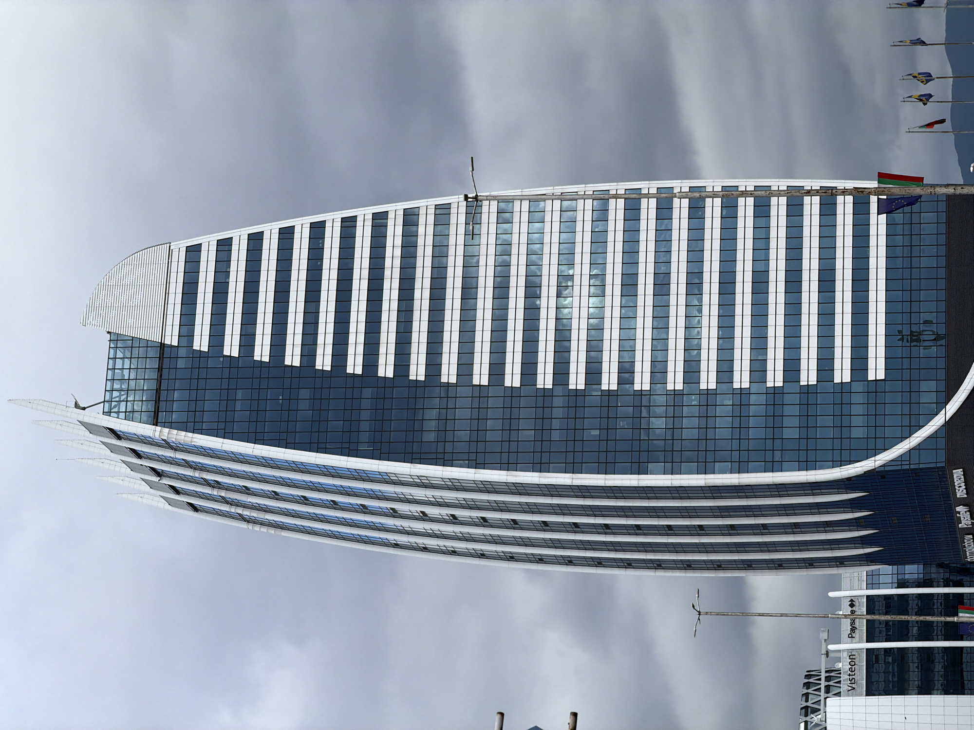 The image features a modern, curved building in Sofia, Bulgaria, likely a commercial or office structure. The building's facade is primarily glass with white vertical accents, creating a striped appearance. Sunlight reflects off the glass, and several flags, including what appears to be the European Union flag, are displayed on poles near the top of the building. The sky is overcast. The building's lower section has a layered, curved design. In the foreground, there's a glimpse of another building with visible company logos such as "Visteon" and partial signs of other companies. A lamppost stands nearby. The environment suggests a business district.