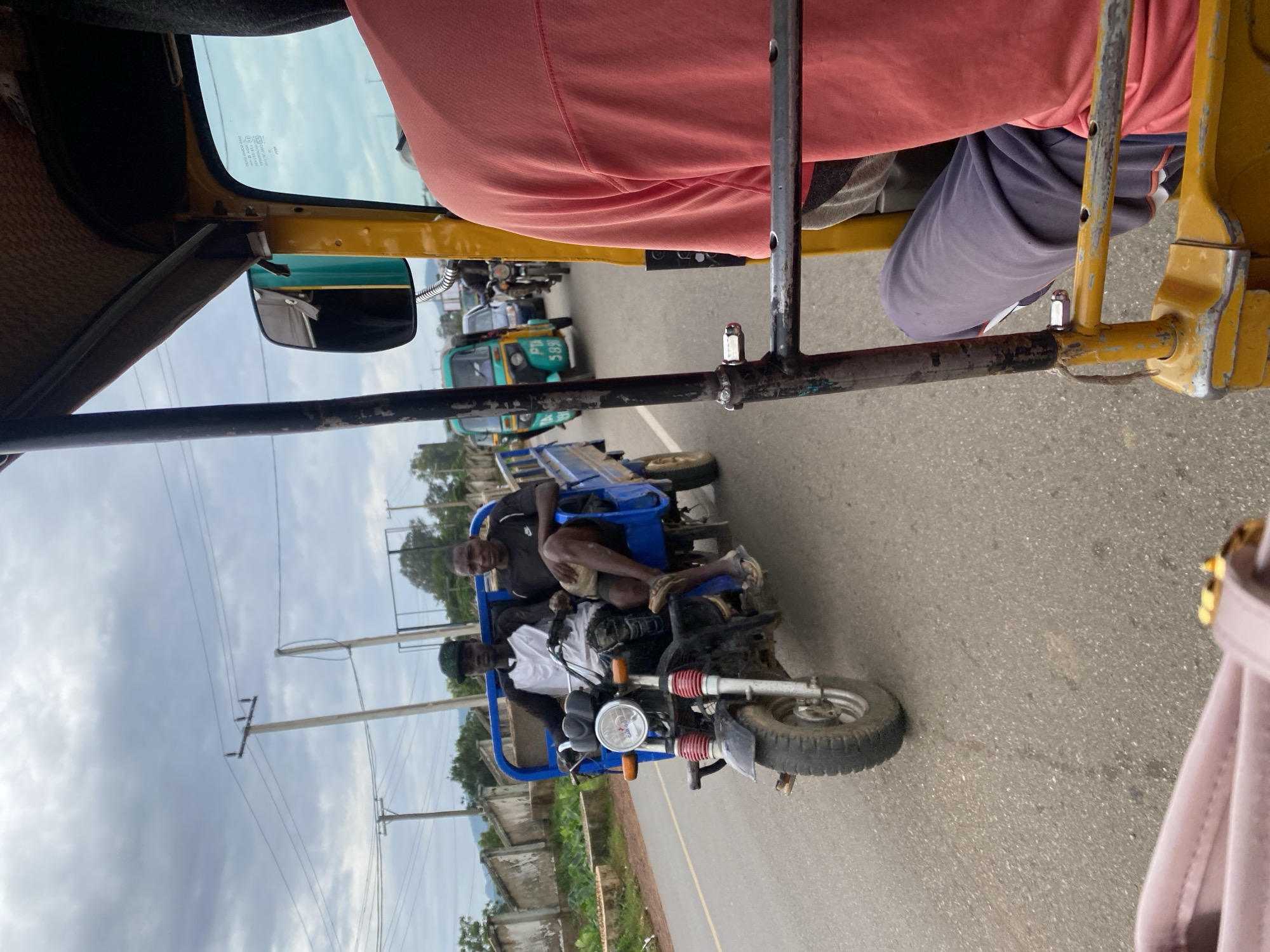 The image captures a scene from a vehicle, likely a trishaw, in motion on a paved road in Rafin Par, Nigeria. The primary subjects are two men riding in the back of a blue trishaw, followed by other vehicles including a green and blue trishaw and motorcycles. The perspective is from within the yellow trishaw, looking forward and to the side. The sky is overcast with gray clouds, suggesting a cloudy or potentially rainy day. The surrounding area shows vegetation and some structures that appear to be buildings with roofs and walls, indicating a semi-urban or rural setting. Power lines are visible, a common sight in many regions. The men in the trishaw appear calm, with one looking forward and the other to the side. One of the men is wearing a dark t-shirt and shorts, while the other wears a white t-shirt and a dark cap. The overall impression is of daily transportation in a Nigerian town. The time of day is not explicitly discernible but the lighting suggests it is daytime. There is no visible text in the image.