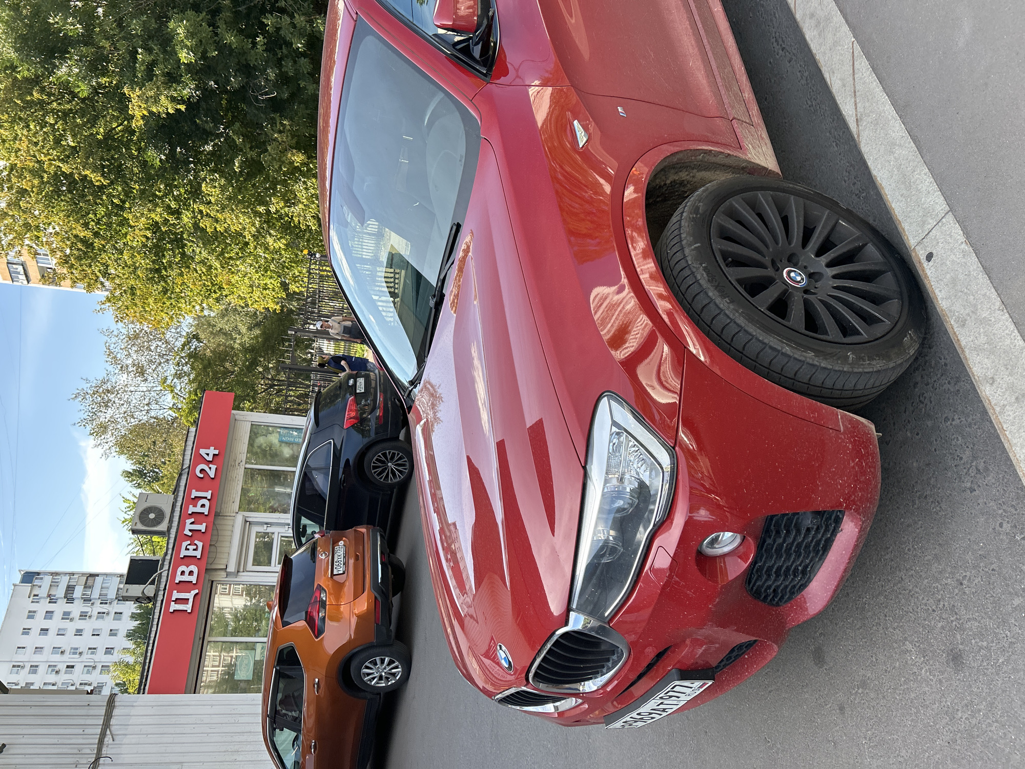 The image shows a red BMW X4 parked on a street in front of a flower shop called "ЦВЕТЫ 24" (Flowers 24). The flower shop is a small building with a red awning and large glass windows. Inside, some flowers are visible. The street is paved with asphalt and has a sidewalk made of concrete and stone. There are other cars parked on the street, including an orange Nissan Qashqai and a black Audi Q5. In the background, there is a tall apartment building and trees. The sky is blue with some clouds. The red BMW is the main subject of the image, and its front is facing the camera. The car has a license plate that reads "E969AT 977". There are two women standing near the black Audi, and one man standing near the fence in the background. The scene appears to be in a residential area of Moscow, Russia, given the architectural style of the buildings and the visible Cyrillic text on the shop's awning and the car's license plate.