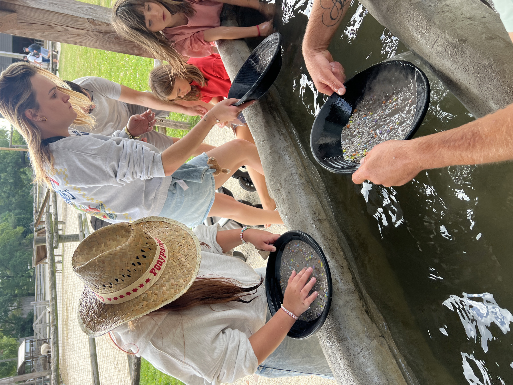 In the city of Collendoorn, Netherlands, a group of individuals are engaged in an activity that appears to be gold panning. The scene unfolds in an outdoor setting, possibly a park or a recreational area, with a long, concrete trough filled with water serving as the primary focal point.
Several people are gathered around the trough, with varying degrees of focus on their tasks. A young girl, wearing a pink t-shirt, is partially visible in the background, her hands submerged in the water. To her left, another young girl with brown hair, dressed in a red t-shirt, is also participating, her hands in the trough.
In the foreground, a person wearing a straw hat with a red band that reads "Ponypark" is holding a black pan filled with water and what appears to be small, colorful pebbles or stones. Their hands are actively sifting through the contents. Beside them, another individual, whose arms and tattooed shoulder are visible, is also holding a similar black pan, tilting it to allow water to flow out, revealing a collection of the small, colorful items. The hands of this individual are positioned as if to carefully separate or inspect the contents of the pan.
The water in the trough is murky and reflects the sky and surrounding greenery. The trough itself is made of concrete and shows signs of wear. Behind the trough, wooden fences and structures can be seen, suggesting a rustic or farm-like environment. In the far background, a sheep is visible near a wooden enclosure, further reinforcing the rural or park-like atmosphere. The overall impression is one of a leisure activity, likely educational or entertaining, taking place in a naturalistic setting.