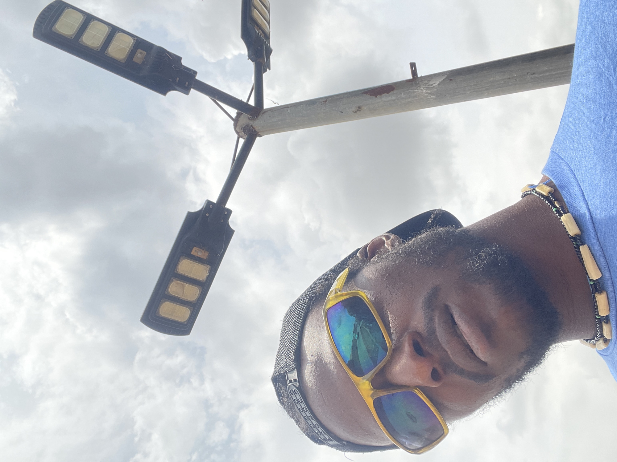 An eye-level, rotated image shows a man facing downward, wearing yellow-framed sunglasses and a black cap, standing beneath a streetlight. The man has dark skin, a beard, and a wooden-bead necklace. The streetlight above him has three rectangular LED lights on each of its arms, mounted on a metal pole. The background is a cloudy sky. The location is likely Abuja, Nigeria.