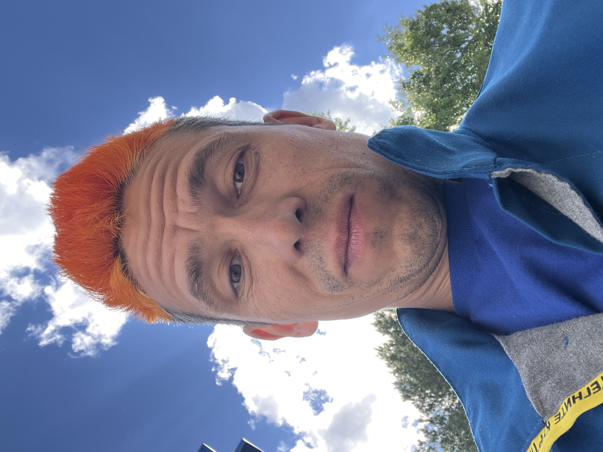 A man with orange hair is looking down at the camera. He has a blue shirt and jacket. The background is a blue sky with white clouds, and a tree. The text on the jacket appears to be in Russian. The image appears to be taken from a low angle, looking up at the man. The location, Chaikovsky, Russia, cannot be verified from this image alone.