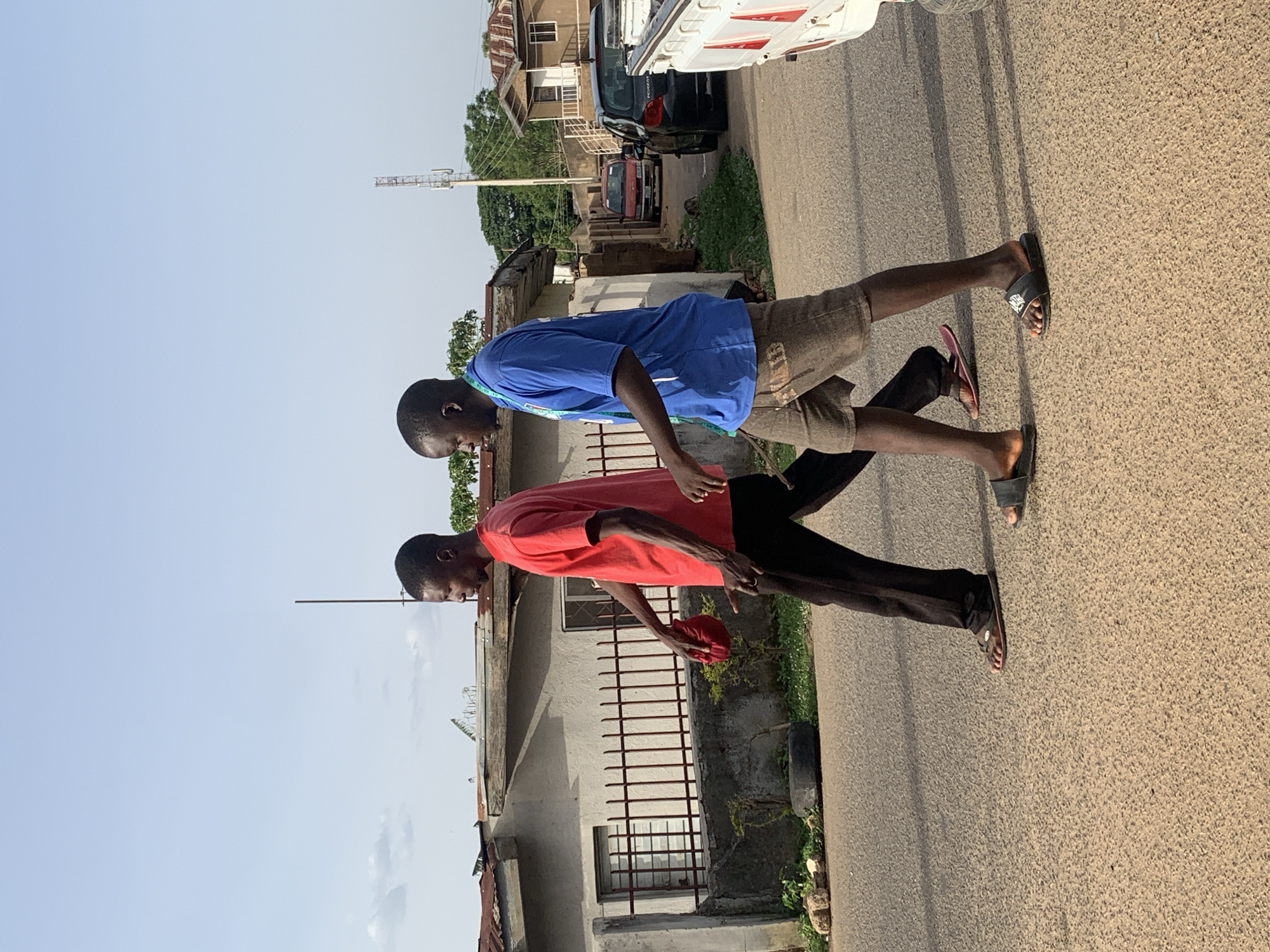 Two young men walk along a paved road. The man in the foreground wears a red shirt and black pants. The man behind him wears a blue shirt and tan shorts. Both are wearing sandals. The location appears to be a residential street in Jos, Nigeria, indicated by the style of buildings and the vegetation. A white truck and other vehicles are parked along the road. The sky is clear.