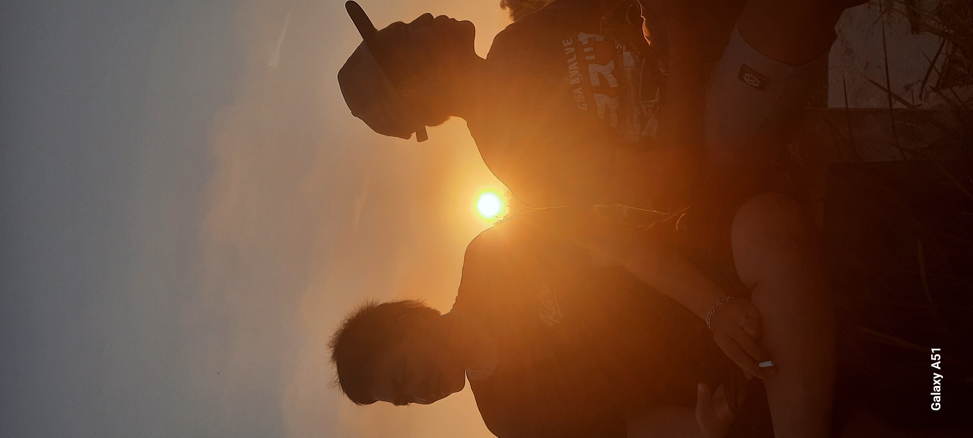 At sunset in Duren, Indonesia, two individuals are silhouetted against the orange sky. The person on the left, with disheveled hair, is holding a lit cigarette and wears a t-shirt with the word "Retro" visible. A silver bracelet adorns their left wrist, and a ring is on their left ring finger. The person on the right wears a baseball cap and a t-shirt with Japanese text and the words "Giga 8 Valve" printed on it. They are also wearing shorts. The scene is set amidst dried grass, and the sun is setting low on the horizon, casting a warm glow over the landscape. The camera's designation, "Galaxy A51," is visible in the bottom left corner.