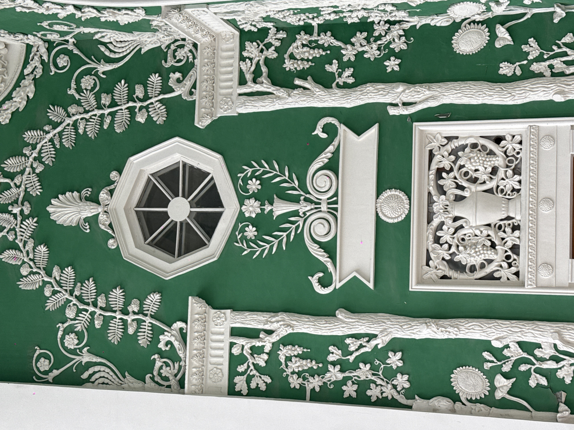 This image shows a detailed section of the ceiling in the Green Dining Room of the Ostankino Palace in Moscow, Russia. The ceiling is predominantly green with ornate white stucco details. These include depictions of floral and plant motifs, such as vines, leaves, flowers, and tree branches. There are also geometric shapes, including an octagonal window or skylight, a rectangular vent with a depiction of grapes and a vase, and a decorative architectural element resembling a banner or column. The stucco is very intricate and three-dimensional.