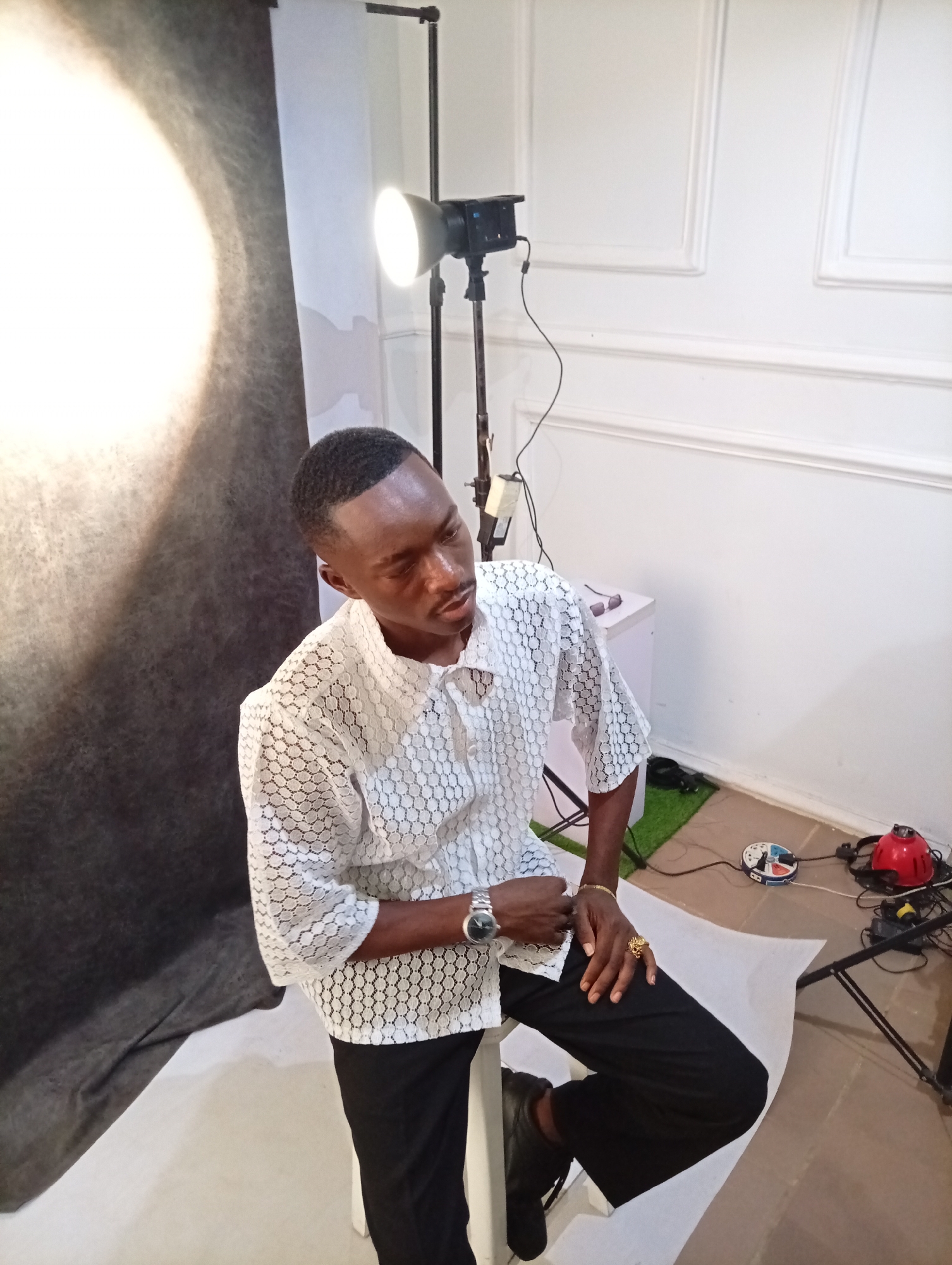 A medium shot depicts a man seated in a studio setting. He is dressed in a white patterned short-sleeved shirt and black trousers, with black shoes peeking out from under his pants. He wears a wristwatch and a ring on his left hand. His hair is closely cropped.
The man is seated on a white object, possibly a stool, and he is positioned against a backdrop that features both a dark gray portion on the left and a white wall with decorative molding on the right. A photographic lighting setup is visible behind him, complete with a stand, light, and attached power cords. On the floor nearby, there is some artificial grass, a white object, a power strip and red object.