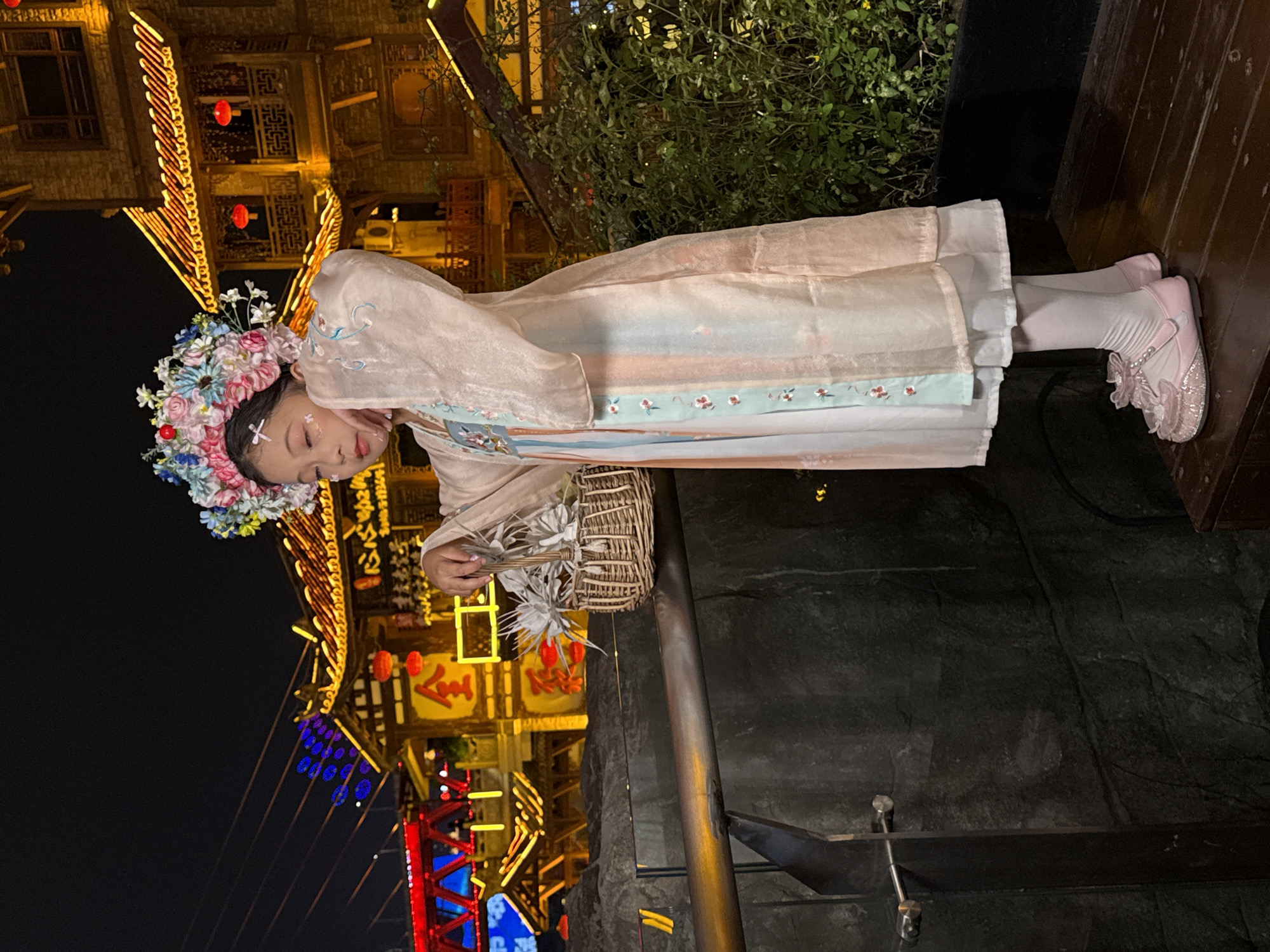 The image features a young girl dressed in an elaborate traditional Chinese costume, likely a form of Hanfu, set against a vibrant night scene. She wears a layered, light pink or beige sheer outer robe over a white dress, which features a light blue band adorned with delicate floral patterns. Her feet are clad in white socks and sparkling pink shoes with floral and ribbon embellishments. Her head is adorned with a large, ornate floral headpiece, predominantly in shades of pink, blue, and white, with small red accents. Glitter or decorative white dots are visible on her face, suggesting festive makeup.
She gently holds a small, intricately woven basket containing white, feathery decorative elements in her hands. Her eyes are softly closed, and her gaze is directed downwards, conveying a serene or contemplative expression, possibly while posing for a photograph.
The background is dominated by traditional Chinese-style buildings, richly illuminated with warm golden strings of lights and hanging red lanterns. Further back, hints of blue and red neon lights suggest a bustling urban environment. Below the girl and to her right, a dark, rocky texture or body of water is visible, separated by a sturdy wooden railing. The scene is clearly set at night under a clear sky, highlighting the intricate lighting of the environment. Indistinct Chinese characters are visible on some of the signs in the background. The overall atmosphere is cultural and festive, characteristic of a special event or performance in a Chinese setting, specifically the Yuzhong District.