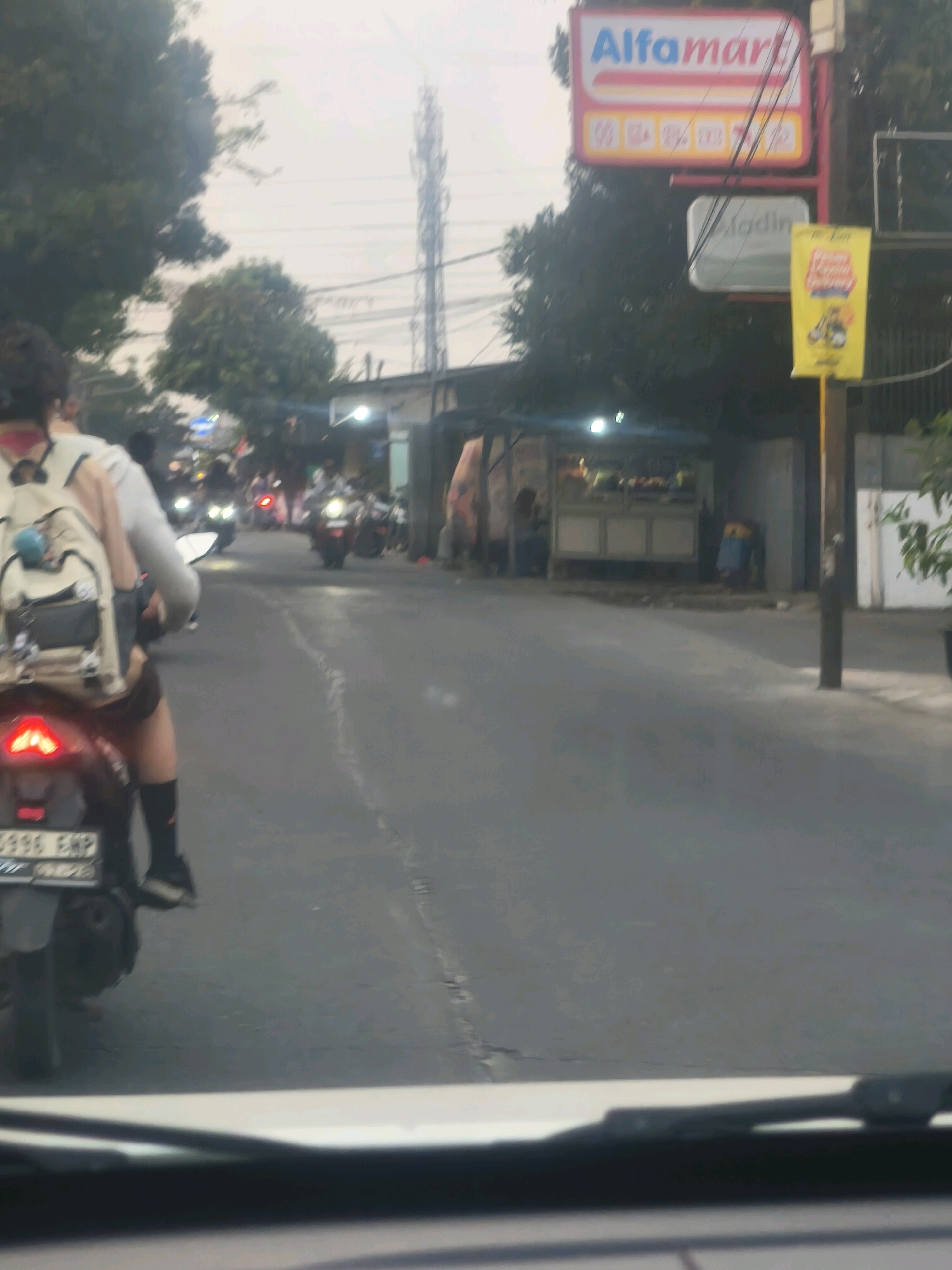 The image depicts a street scene in Bekasi, Indonesia, under an overcast sky. In the foreground, a person rides a motorcycle, viewed from behind. They are wearing a light-colored shirt, dark shorts, black socks, and white shoes. A beige backpack with grey accents is on their back. The motorcycle has a license plate that reads "6996 EMP".
Further down the street, other motorcycles are visible, some with their headlights on, indicating traffic. A prominent red and white sign for "Alfamart" is seen on the right, along with another smaller sign below it. A yellow banner with an image of a motorcycle is also displayed.
On the left side of the street, there appears to be a food stall or small shop with people visible inside and around it. Trees are present in the background, along with a tall communication tower. The road is paved and shows some wear and tear. The overall impression is a typical urban street scene in Indonesia, likely during the daytime given the ambient light.