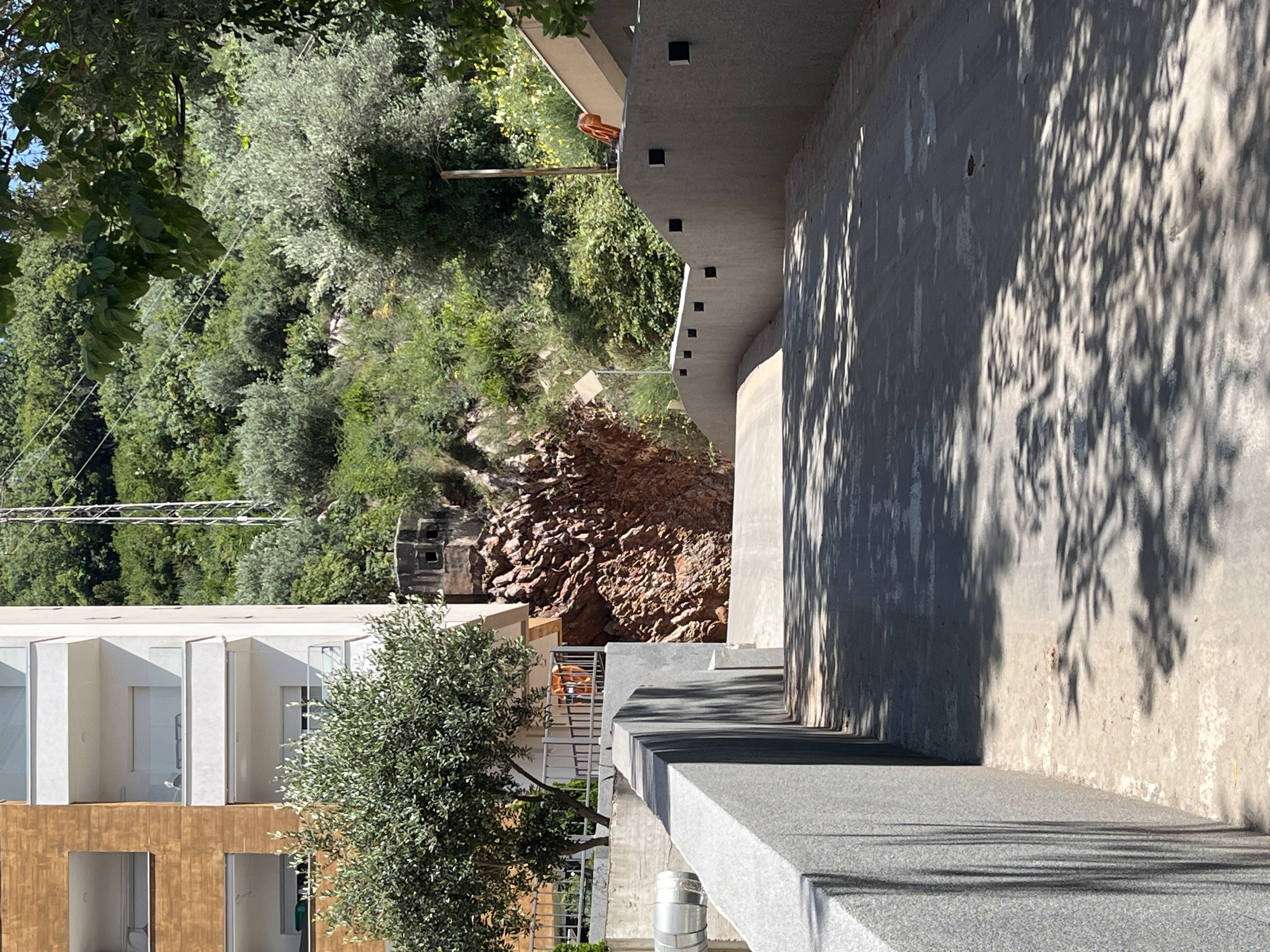 An eye-level outdoor, medium shot captures a paved pathway leading towards a rocky hillside, with a modern building on the left. The pathway is a gentle upward slope with a textured, grey concrete wall on the left and a slightly curved, concrete wall on the right. The wall on the right has small, dark square indentations at regular intervals. The pathway is dappled with sunlight and shadows from overhead foliage.
On the left, the modern building has a rust-colored facade with several levels of balconies, some with glass railings. An olive tree with dense green foliage stands in front of the building, partially obscuring it. Behind the building, a metal fence encloses an area where an orange life preserver is visible. To the right of the building, a steep, reddish-brown rock face is visible. On top of the rock face, a small, concrete structure with two square openings is perched, resembling a bunker.
The background is dominated by a lush, green hillside covered in trees and vegetation. A tall, metallic tower structure with crossbeams stands on the upper right portion of the hillside. The sky is bright blue, suggesting a sunny day. The scene appears to be located in Budva, Montenegro, possibly in a residential or urban area adjacent to a natural landscape. There are no people or animals visible in the frame.