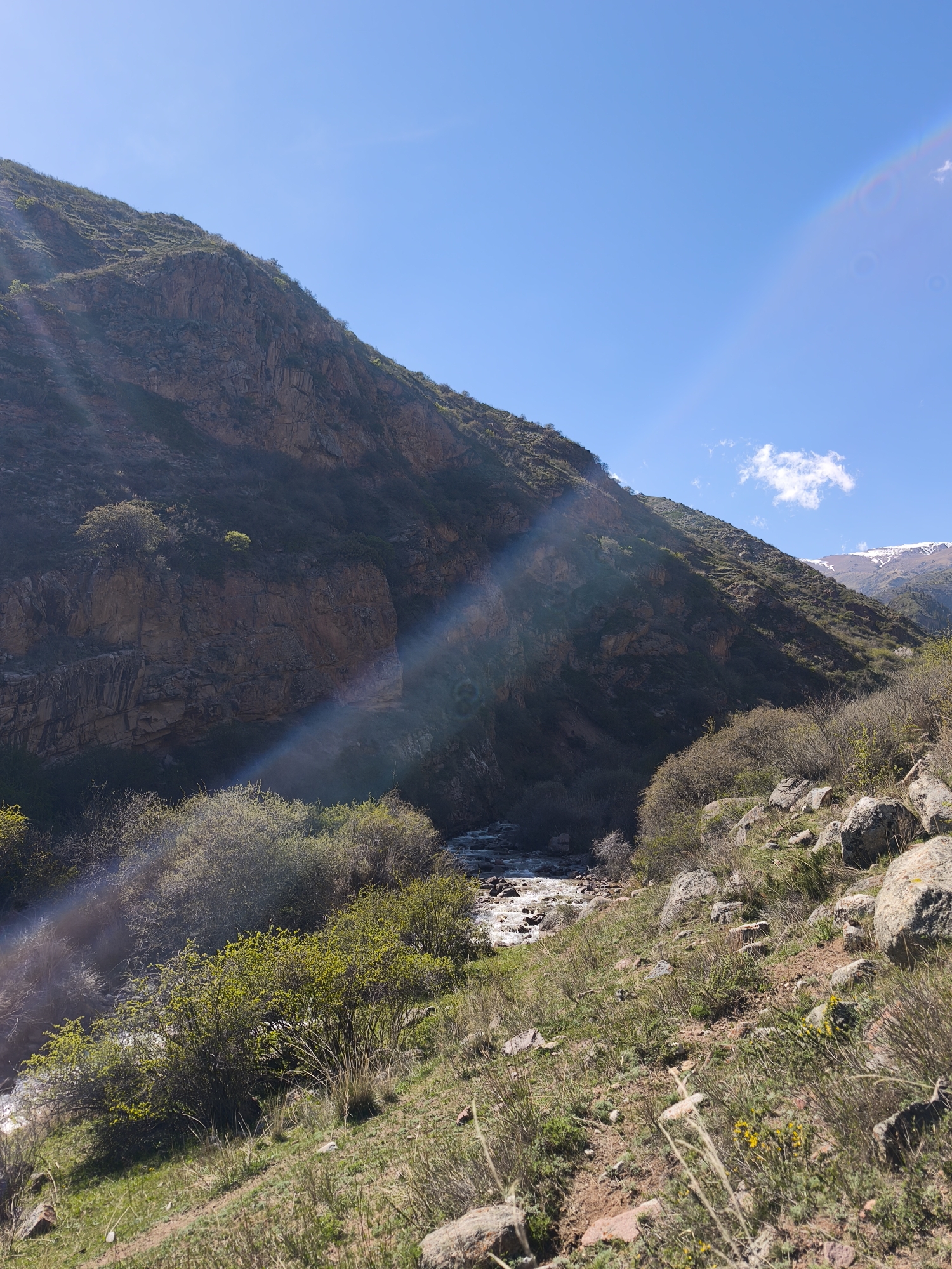 The image shows a mountain landscape in Cholpon Ata, Kyrgyzstan. A mountain occupies the left side of the frame, its surface covered in rock formations and sparse vegetation. Sunlight filters through the scene, creating lens flare. At the base of the mountain, there is a river. On the right side of the frame, there is a grassy hillside with scattered rocks and bushes. In the distance, snow-capped mountains are visible beneath a blue sky with a few clouds.