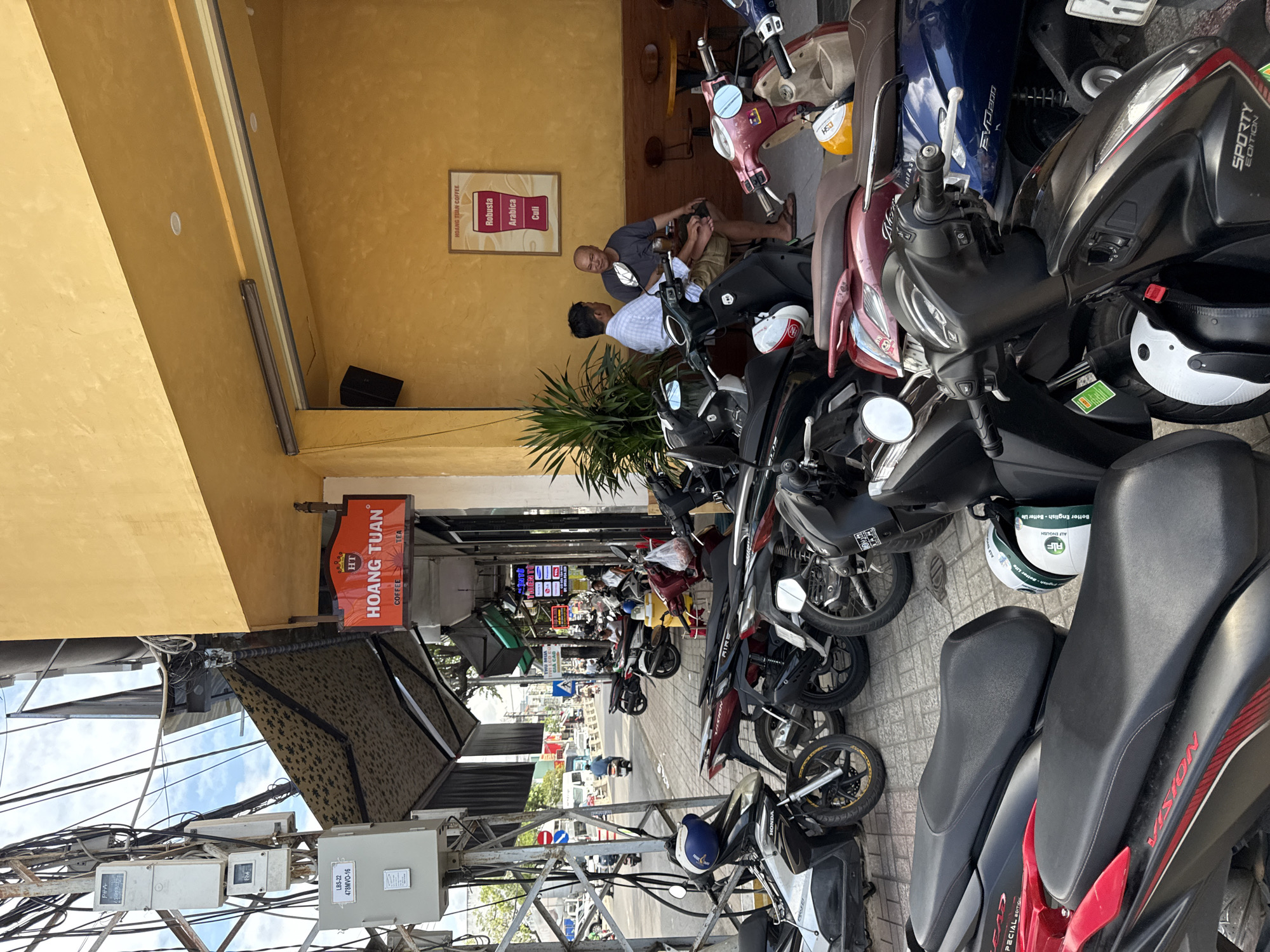 The image depicts a street scene in Nha Trang, Vietnam, likely outside a cafe or shop. A row of parked motorbikes, predominantly scooters, occupies the foreground and sidewalk. The motorbikes vary in color and style, with some showing wear and tear. Two men are seated at a table outside a building with a bright yellow facade. One man, wearing a blue and white checkered shirt, is facing away from the camera and appears to be talking to the other man, who is wearing a grey t-shirt and shorts and is looking towards the first man. A framed poster advertising coffee is visible on the yellow wall behind them. Above the seating area, a black awning extends outwards. Electrical boxes and numerous wires are mounted on a metal pole to the left of the frame. The street in the background is busy with traffic, including more motorbikes and cars. The overall impression is a typical urban scene in Vietnam, characterized by the presence of motorbikes and outdoor seating for establishments.