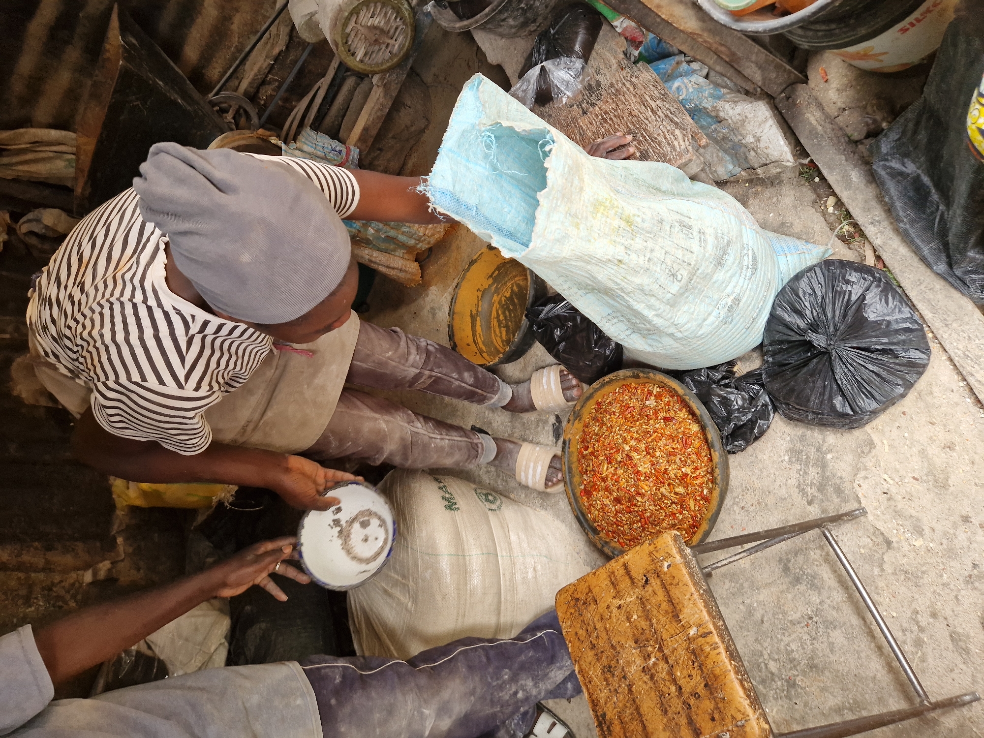 In Kabong, Nigeria, an outdoor marketplace scene is depicted with two individuals engaged in an activity. One person, wearing a grey beanie, striped shirt, and brown pants, leans over a pile of red, chopped chili peppers in a large black bowl. They are holding a small white bowl with some debris and are reaching towards a large, light blue woven bag, which appears to be an open sack. Another person, partially visible on the left, wears a grey t-shirt and blue pants and is also reaching towards the white bowl. The ground is concrete, and the background shows a rough, shed-like structure with corrugated metal roofing and wooden planks. Various other items are scattered around, including a wooden stool, a yellow bowl, black plastic bags, and other containers.