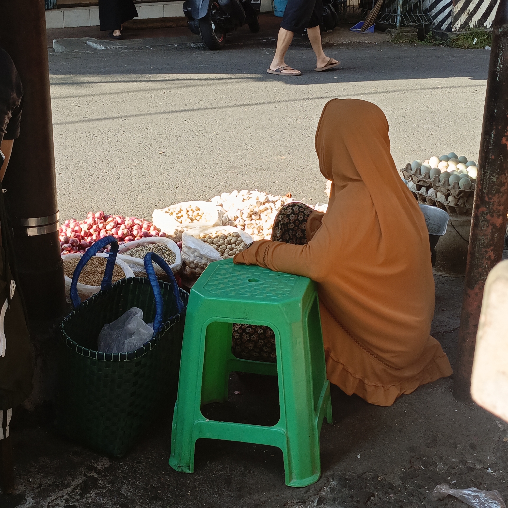 An overhead view captures a vendor kneeling on the ground beside a bright green plastic stool, dressed in an orange hijab and robe. They are surrounded by an array of produce including sacks of grains and piled red onions. Two woven baskets, one dark green with blue trim and the other a lighter green, sit nearby, holding what appears to be plastic bags. In the background, a rack of eggs is visible. The scene is set on a paved street in Salatiga, Indonesia, with glimpses of passing pedestrians and a motorcycle. The lighting suggests it is daytime, with shadows cast by the overhead structure.