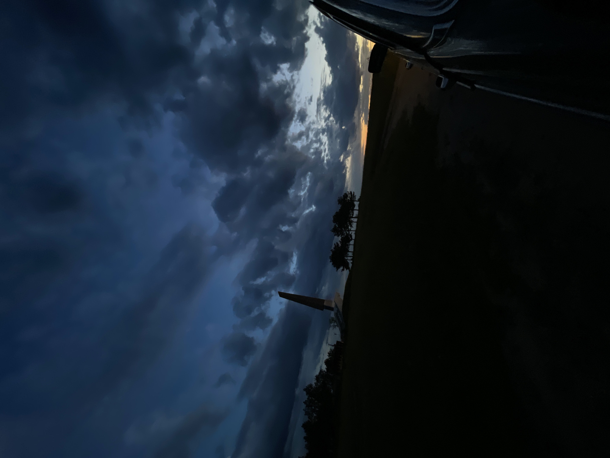 The image shows a low-angle view of the Bender Fortress Memorial in Kitskany, Moldova, taken from a vehicle. The memorial, consisting of a tall, pointed obelisk and several rectangular bases, is silhouetted against a dramatic, cloud-filled sky at twilight. The foreground is dark, with a field and trees. A portion of the vehicle from which the photo was taken is visible in the upper-right corner.