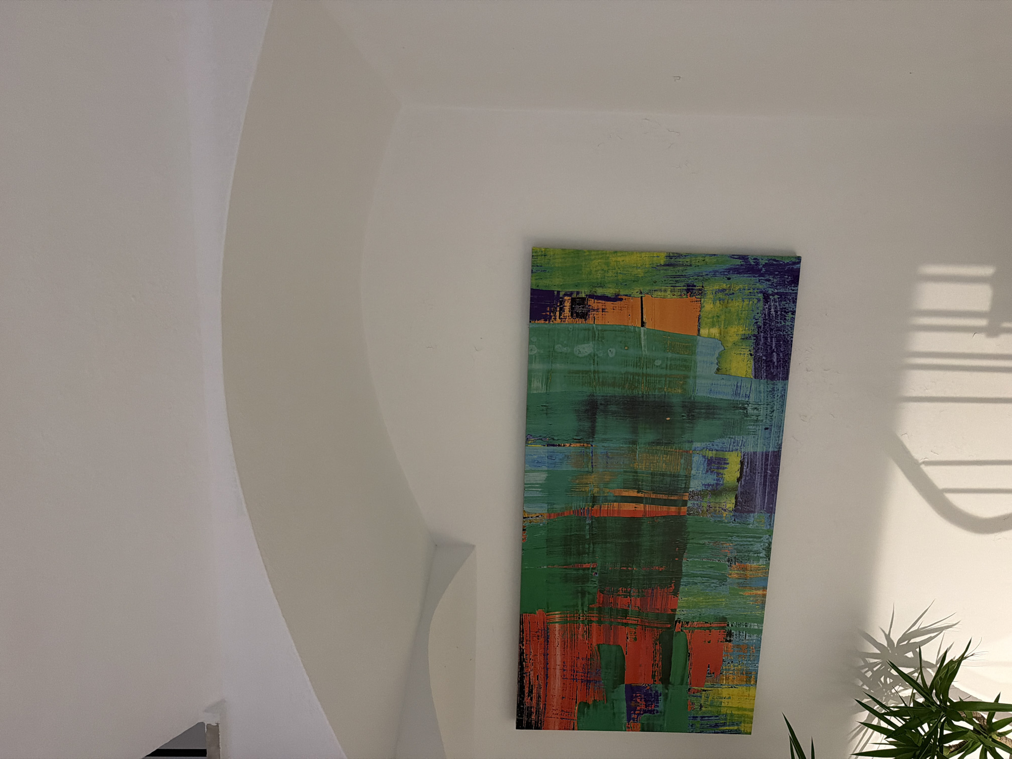 This is a vertical shot of a modern, abstract painting hanging on a white wall. The painting is rectangular and features broad, textured strokes of various colors, including shades of green, orange, yellow, blue, and purple. The artistic style suggests a contemporary abstract expressionist approach. To the left of the painting, a smooth, curved architectural element, likely part of a staircase or wall feature, casts a soft shadow. To the right of the painting, a window casts a distinct shadow of horizontal bars onto the wall, suggesting it's daytime and sunlight is filtering through. In the lower right corner, the spiky leaves of a green plant are visible, casting their own shadow on the wall, further indicating natural light. The overall scene conveys a sense of modern interior design, possibly in a home or gallery setting. The location context suggests it is in Sant Antoni De Portmany, Spain. There is no visible text.