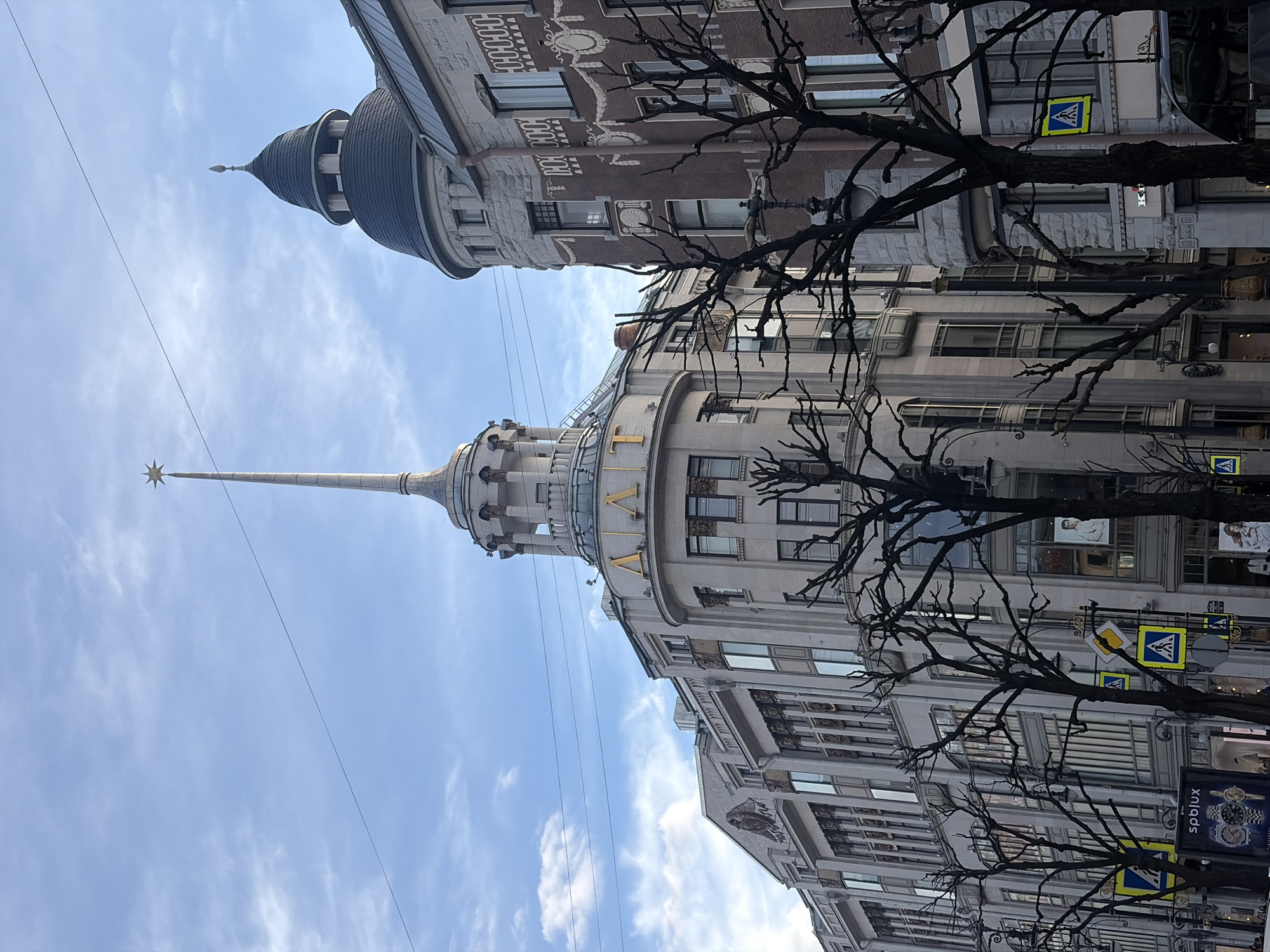The image shows two prominent buildings in Saint Petersburg, Russia, taken from a street-level perspective, rotated 90 degrees counterclockwise.
In the foreground, the Singer House (also known as the Dom Knigi, or House of Books) is prominently featured. The building is topped with a large glass dome and a spire with a star. Large gold Cyrillic letters, potentially "ДОМ" or "КНИГ", are visible below the dome.
Behind and slightly to the left, another building with a turret-like structure is visible. This building appears to be of a different architectural style than the Singer House.
Bare tree branches frame the view, obscuring parts of the buildings and adding depth to the image. Street signs with pedestrian crossings are attached to poles, as well as a wristwatch billboard. The sky is pale blue with scattered white clouds. Electrical wires run across the top portion of the frame.