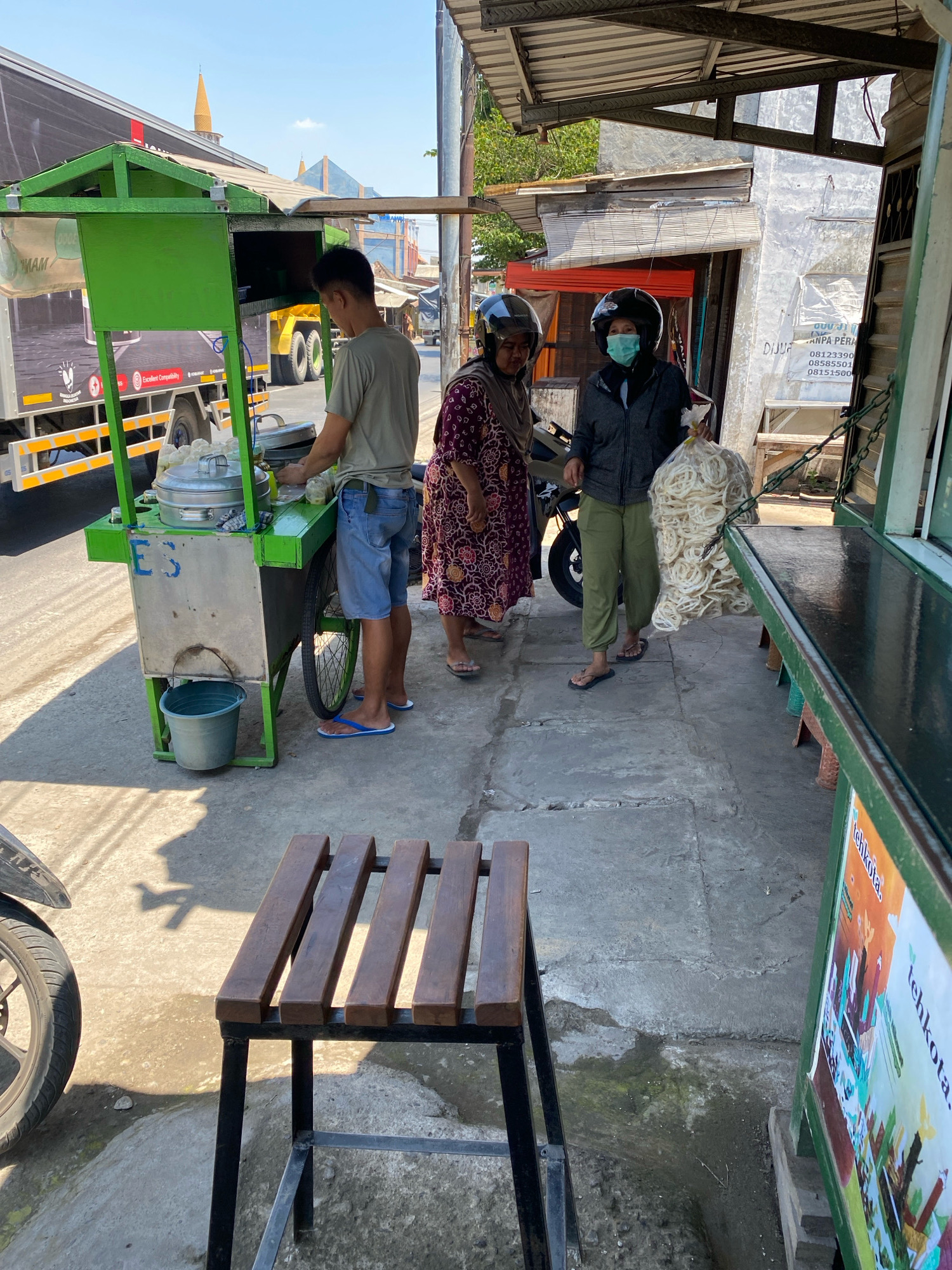 In Perning, Indonesia, a street vendor prepares food at a bright green cart on a sunny day. The vendor, a man wearing a grey t-shirt and blue shorts, stands behind the cart, which displays a metal food steamer. Two women, one wearing a patterned dress and the other a grey jacket, stand near the cart. The woman in the grey jacket wears a light blue mask and holds a large plastic bag filled with white, circular snacks. A motorcycle is parked to the left of the frame. The ground is paved with concrete, and a wooden bench sits in the foreground. Buildings with weathered walls and corrugated metal roofs line the street in the background, suggesting a commercial or residential area. A large truck is parked to the left of the scene, partially obscuring some of the buildings. The overall impression is of a typical street scene in a bustling Indonesian town.