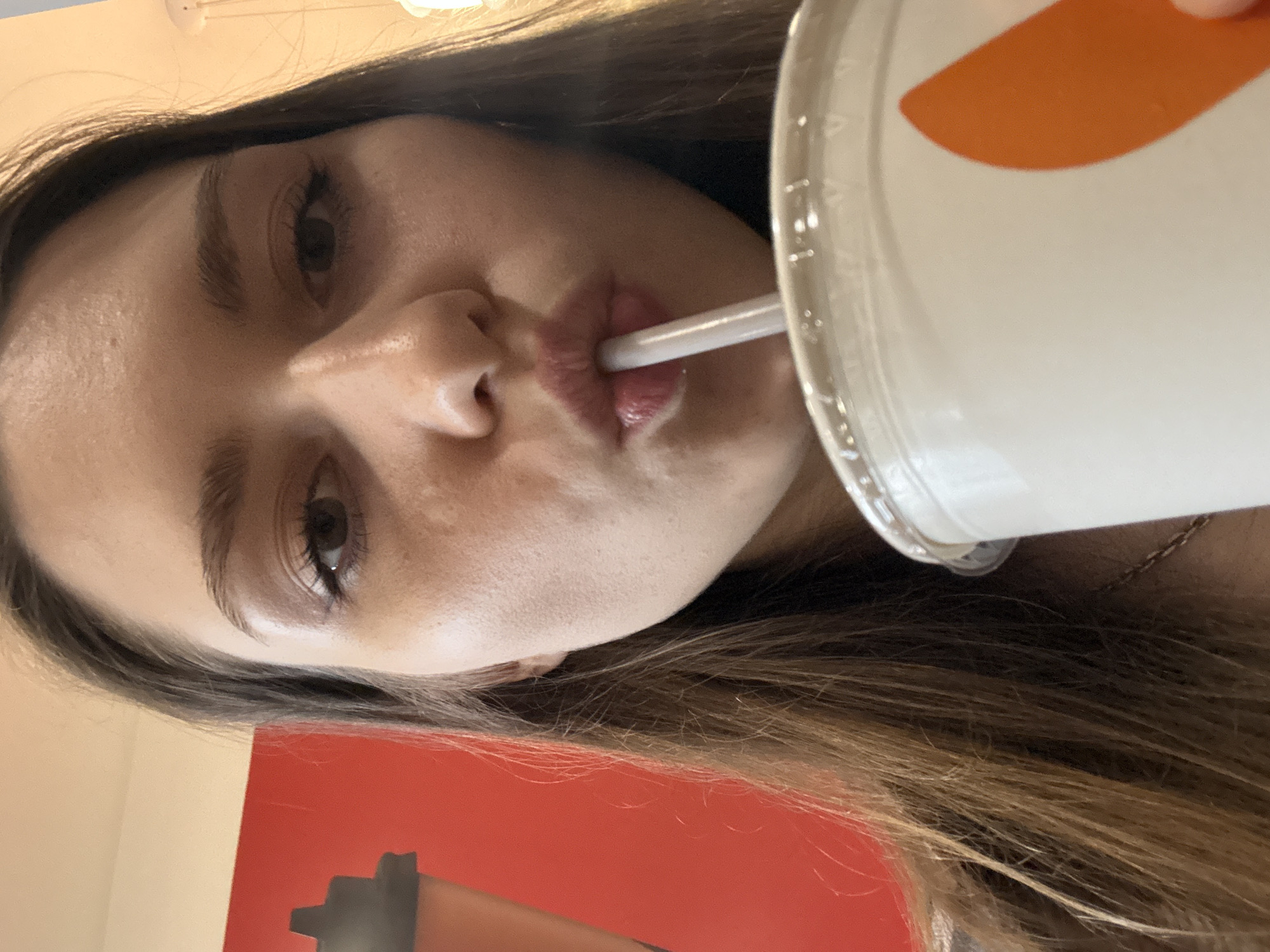 The image, rotated counter-clockwise by 90 degrees, presents a close-up of a young woman engaged in drinking from a large cup through a straw. Her face is angled towards the viewer, with her dark eyes looking towards the camera. Her long, dark brown hair frames her face and extends across the lower portion of the frame.
The woman's lips are pursed around a white straw, which is inserted into a tall, white, cylindrical cup topped with a clear plastic lid. A distinctive orange, curved design or logo is visible on the side of the cup. Her facial expression is neutral to slightly focused on the act of drinking. A delicate necklace chain is subtly visible around her neck.
The setting appears to be indoors, under artificial lighting. The background consists primarily of a plain red wall, with a lighter-colored wall above it and a light fixture partially visible at the top. An indistinct, dark object can be seen on the red wall. The activity is the woman drinking from a beverage container. The location context is Saint Petersburg, Russia.