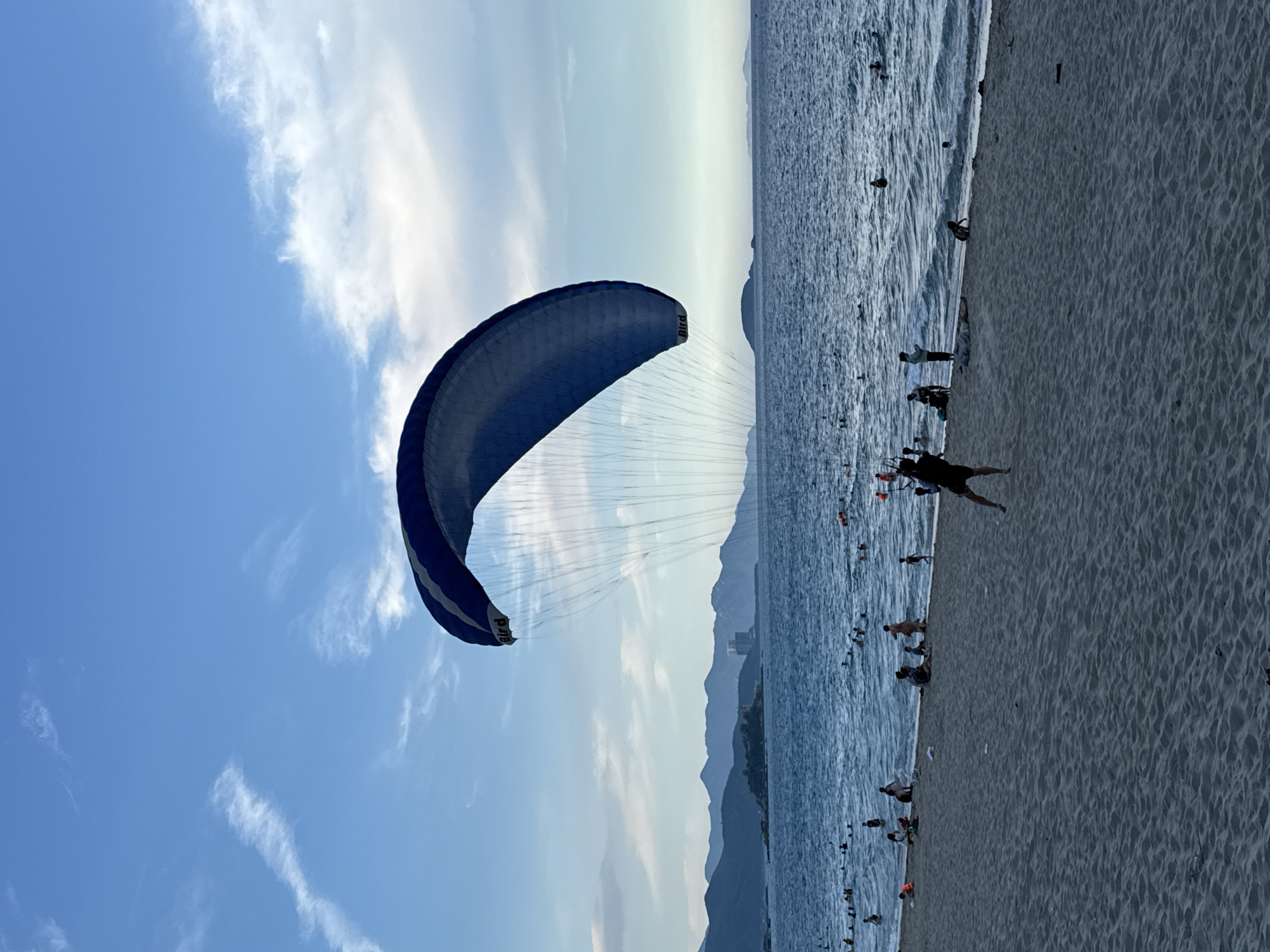 A parasailer with a blue and white canopy is in the air over a crowded beach and ocean. The parasailer is wearing dark clothing. Many people are in the water and on the sand. The ocean has moderate waves, and the sky is blue with some white clouds and wispy contrails. In the distance, there are mountains and a few buildings. The beach is made of sand and appears to be a popular recreational area in Nha Trang Ward, Vietnam.