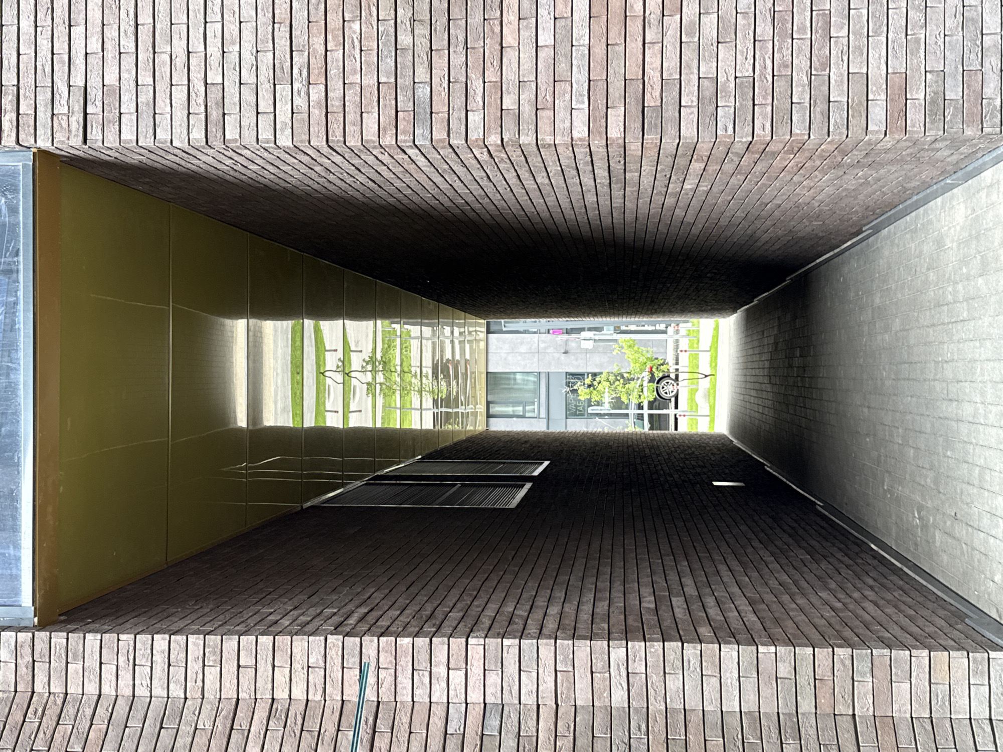 The image depicts a long, rectangular architectural passage or tunnel, viewed head-on and looking directly through its length towards a brightly lit exterior at the far end.
The passage is constructed from four distinct surfaces:
The **left and right walls** of the passage are formed by the reddish-brown brickwork prominently featured in the top and bottom sections of the image. These bricks are laid vertically in a stretcher bond pattern, creating a textured surface, and they recede into the distance, defining the side boundaries of the tunnel.
The **ceiling** of the passage is the smooth, highly reflective olive-green or bronze-colored paneling on the left side of the interior. This surface vividly reflects the outdoor environment beyond the passage, showing bright green foliage (likely trees and grass), elements of modern buildings, and a partial view of a vehicle wheel. Faint, distorted white text, possibly reading "VAPO TAG," is discernible in one of the reflections.
The **floor** of the passage is the dark grey brick surface on the right side of the interior. It appears somewhat smoother than the side walls and is largely obscured by shadow. Two dark, rectangular grates or vents are visible on this surface, located closer to the reflective ceiling.
The interior of the passage is predominantly dark and shadowed, creating a sense of deep perspective that contrasts sharply with the vibrant natural light emanating from the exit. No individuals or specific activities are visible in the scene. The strong exterior light indicates it is daytime. A thin green rod or pole is visible against the bottom reddish-brown brick wall. The location context for this architectural structure is Moscow, Russia.