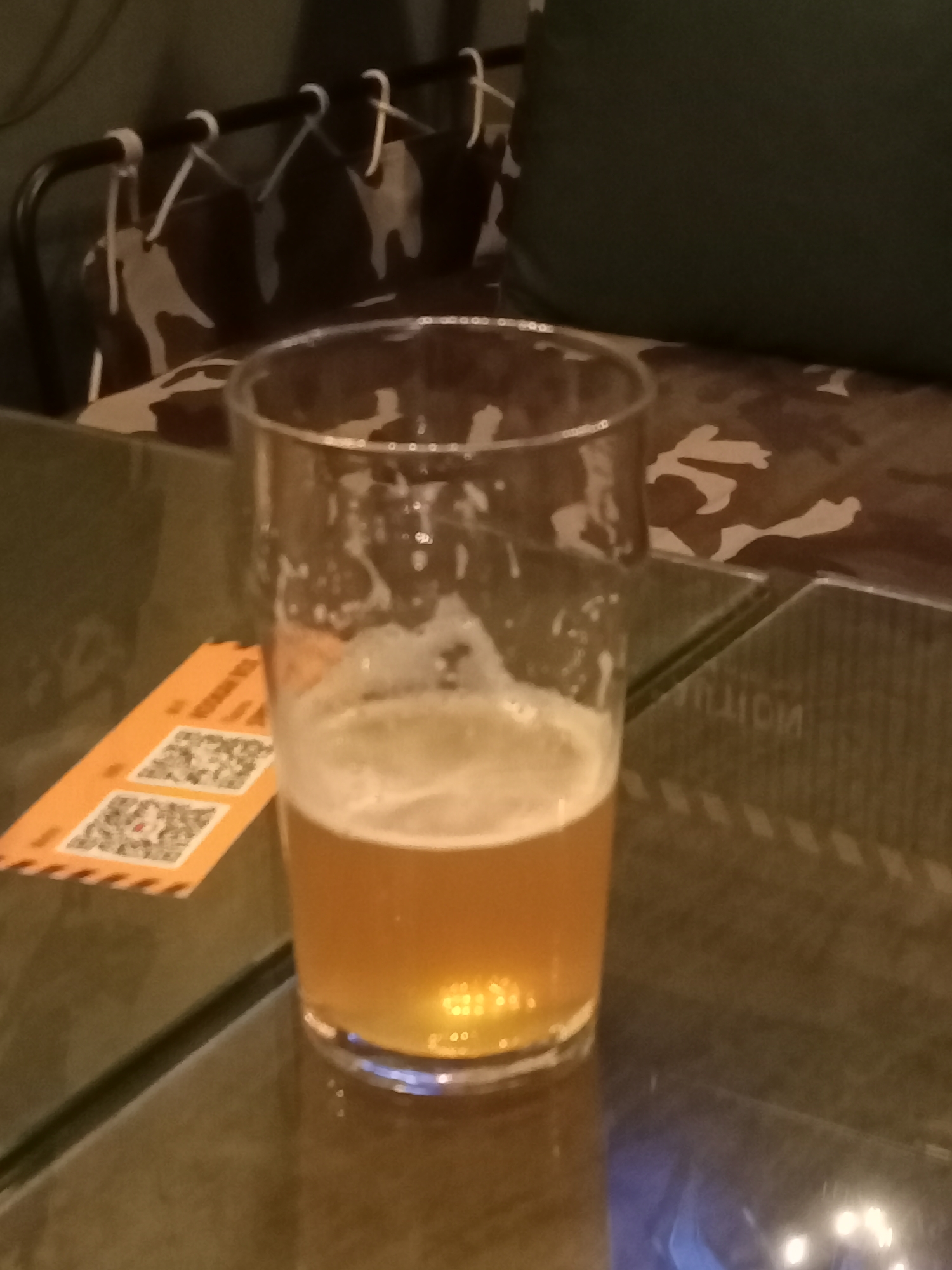 A half-full glass of amber-colored beer with foam rests on a dark, reflective surface. The glass is positioned on the right side of the frame. To the left of the glass, an orange ticket with two QR codes is partially visible. The background features a dark, patterned fabric with camouflage-like shapes and a dark green cushioned surface. A black metal rack with white ties is visible in the upper left corner. The overall lighting is dim, with reflections on the table surface suggesting ambient light sources. The scene appears to be indoors, possibly in a bar or restaurant. No specific landmarks indicating Saint Petersburg, Russia, are discernible.