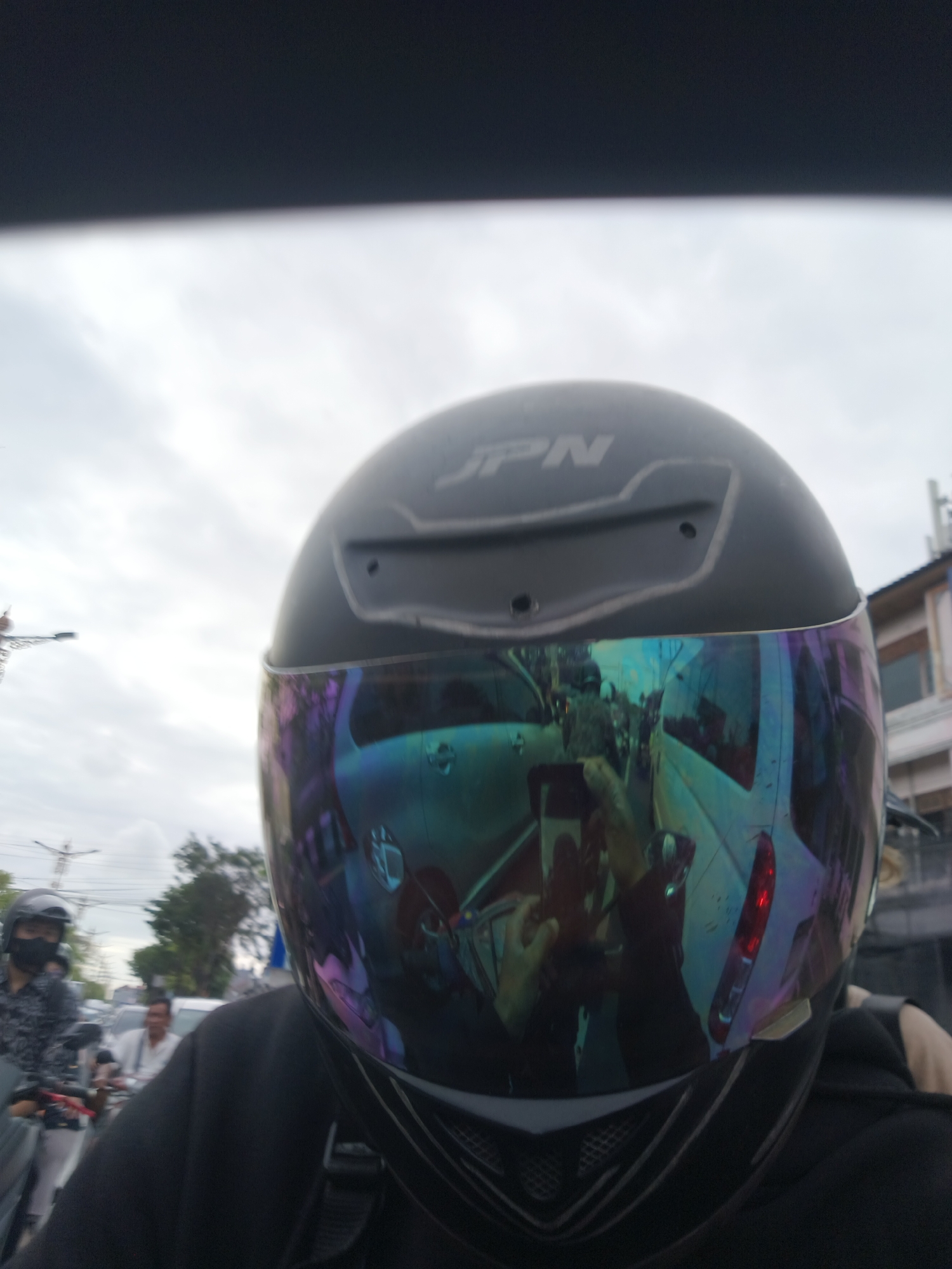 The image shows a first-person perspective of someone wearing a black motorcycle helmet with a reflective visor. The visor displays a distorted reflection of traffic, including cars and a van, suggesting the subject is on a road, possibly in a city or town. The sky appears overcast, indicating a cloudy day.
In the reflection on the visor, another person is visible wearing a black mask and a patterned shirt, also on a motorcycle. The person in the foreground appears to be wearing a black hoodie. The overall scene depicts a moment of travel in what seems to be a busy environment.
The helmet has "JPN" written in white on the top. The background shows some buildings and trees, with streetlights visible in the distance. The lighting suggests it is daytime, but the cloudy conditions create a diffused light. No specific emotions are evident, other than the typical focus and awareness associated with riding a motorcycle in traffic. The location context provided is Kuta Selatan, Indonesia.