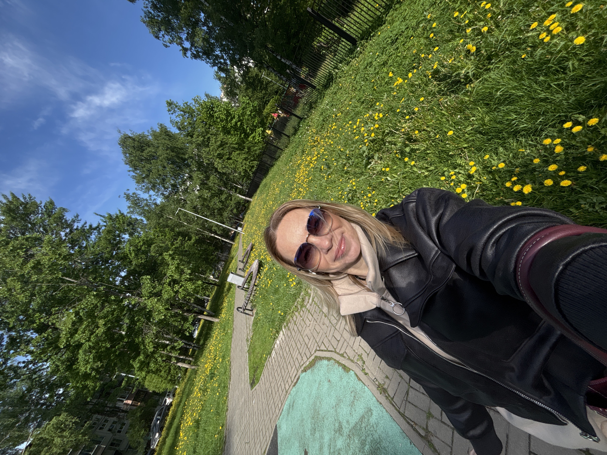 A selfie taken outdoors at a park in Saint Petersburg, Russia on a sunny day. A woman wearing sunglasses and a black jacket poses in front of a field of dandelions, which is surrounded by trees. A paved path with park benches is visible in the background. The sky is blue with some scattered clouds. A black fence is visible in the midground.