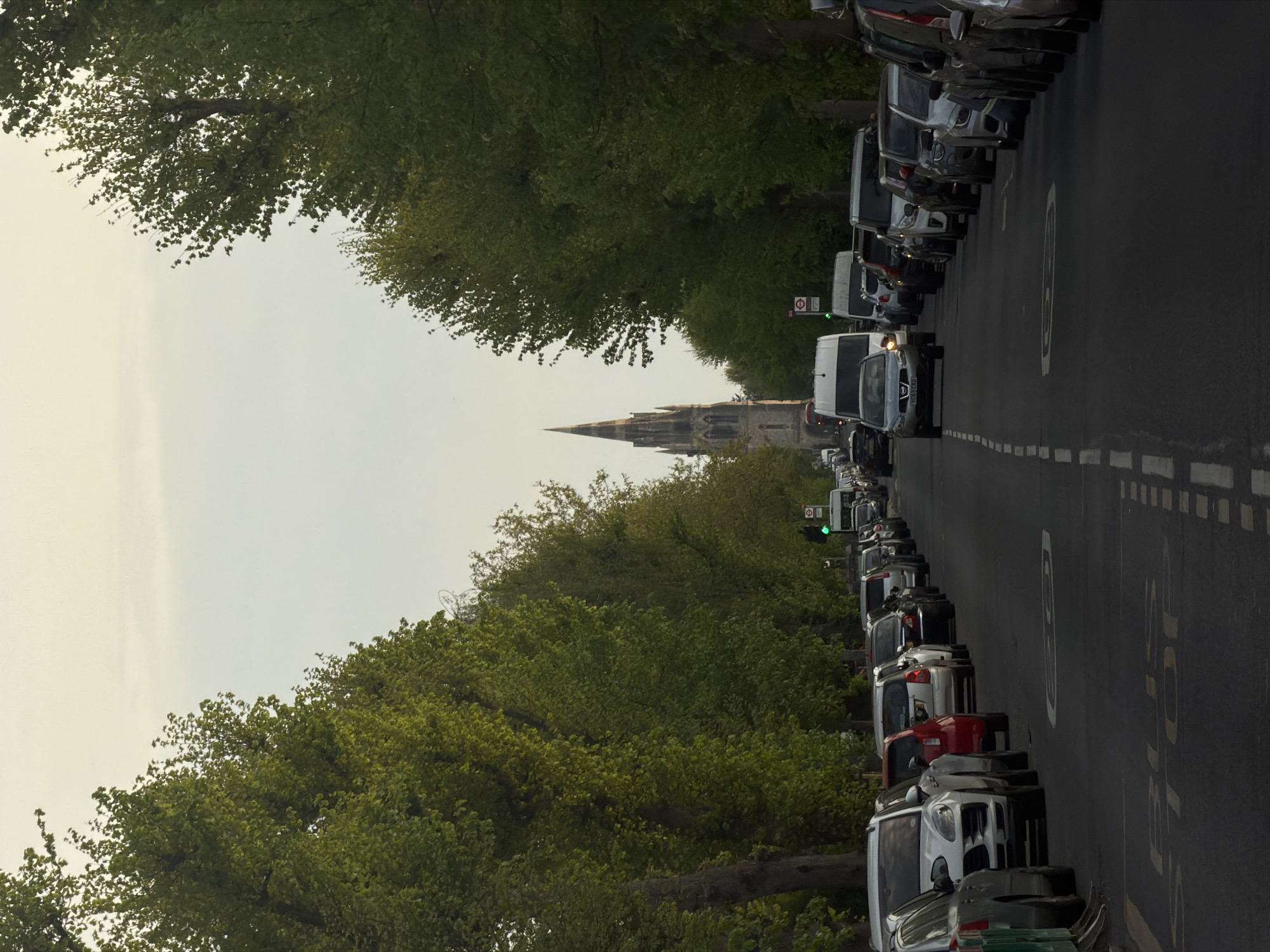 A road in London, United Kingdom, is lined with parked cars on both sides. A line of cars travels down the middle of the road towards a church steeple in the distance. Green trees line both sides of the street. The sky is overcast.