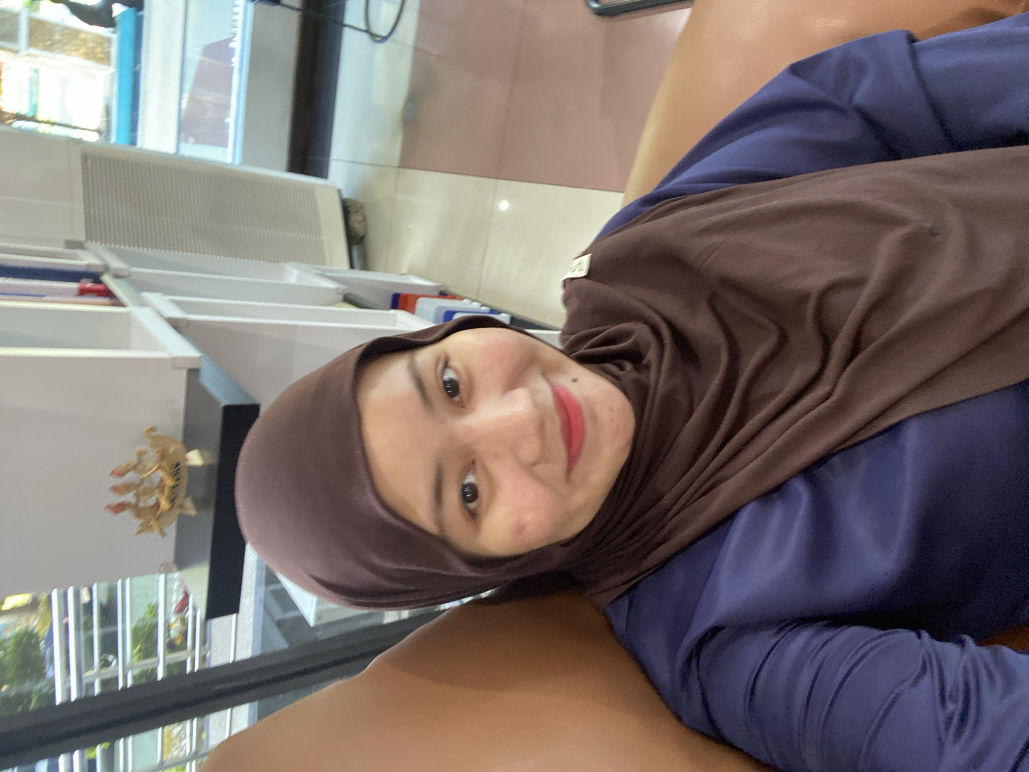 A young woman wearing a brown hijab and a blue shirt is seated in a tan leather chair, facing the camera. She has fair skin, dark eyes, and is wearing red lipstick. The background shows a bookshelf with some books, a decorative ship figurine, and a tall white appliance that appears to be an air cooler or dehumidifier. Natural light streams in from a window behind her. The setting appears to be an indoor commercial space, possibly a cafe or a shop, in Banjarbaru, Indonesia, given the architectural style and the presence of an air cooler, which is common in humid climates.