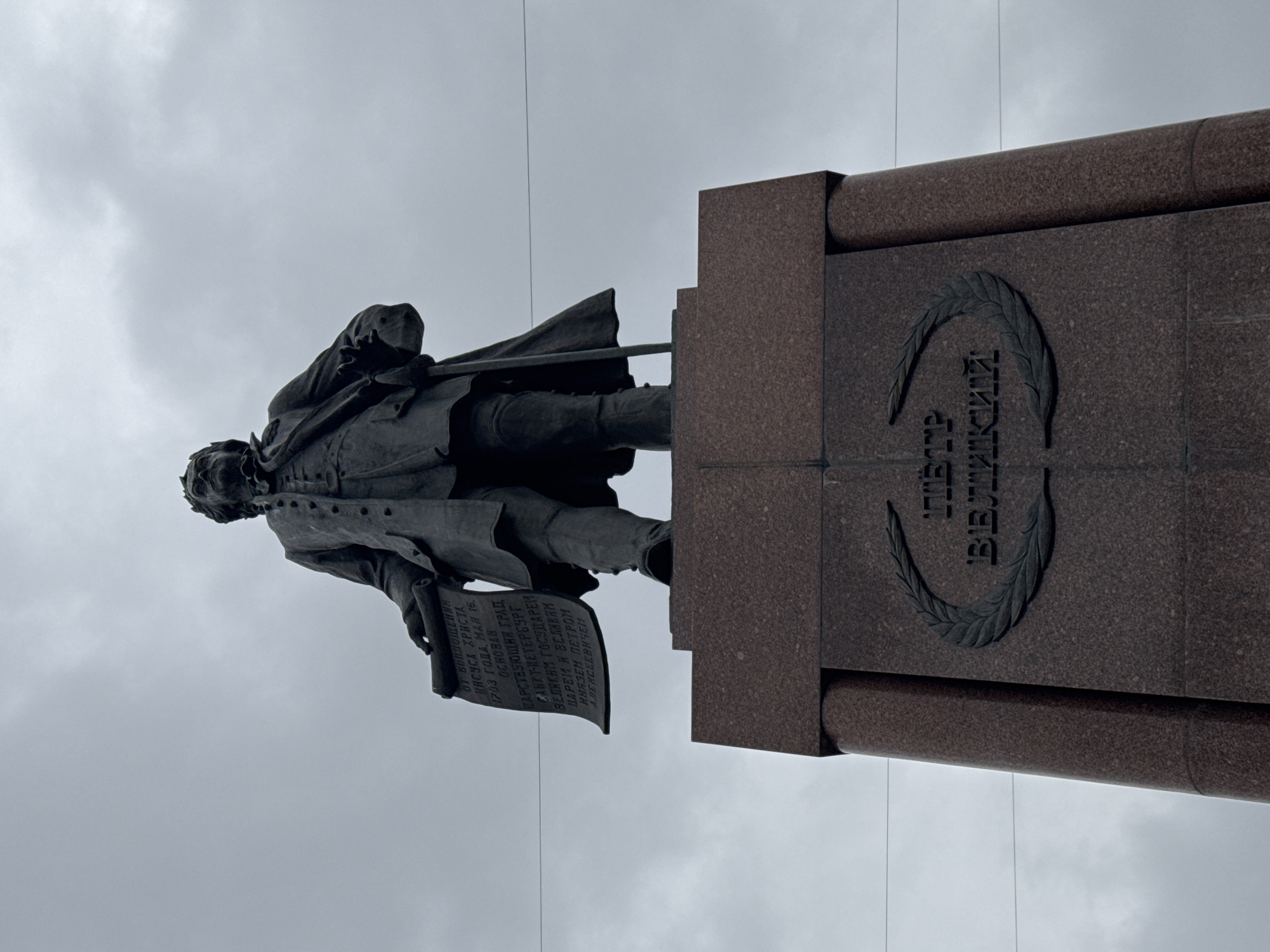The image shows the Peter the Great Monument, a statue of the Russian Tsar Peter the Great, in Saint Petersburg, Russia. The statue is mounted on a tall granite pedestal featuring inscriptions. The sky is overcast.