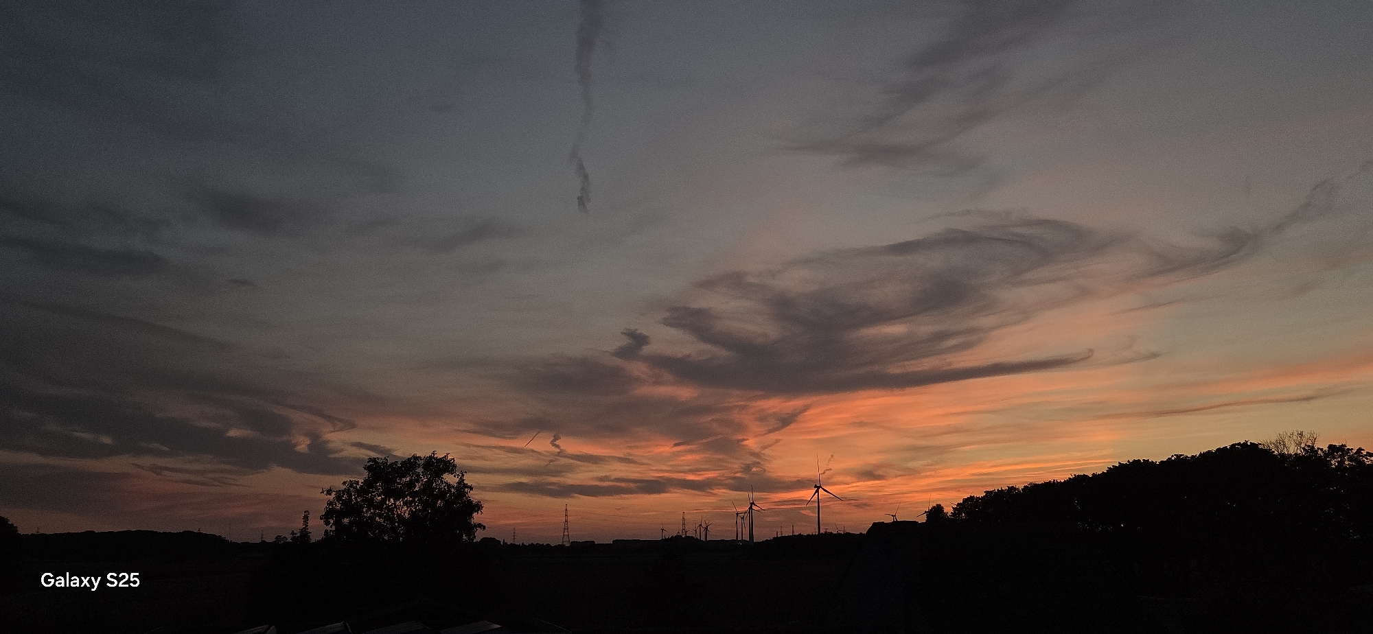 A wide-angle, low-light photograph captures a dramatic sunset over a rural landscape in Eeklo, Belgium. The sky is a canvas of fiery oranges and soft pinks blending into a deep, dusky grey, streaked with wispy clouds. The lower portion of the image is silhouetted against the vibrant sky, revealing a line of wind turbines standing tall, their blades still. In the foreground, a solitary tree with a rounded canopy is silhouetted on the left. Power lines and pylons are faintly visible in the distance. The text "Galaxy S25" is subtly placed in the bottom left corner, indicating the device used for capture. The overall impression is one of serene, natural beauty transitioning into twilight.