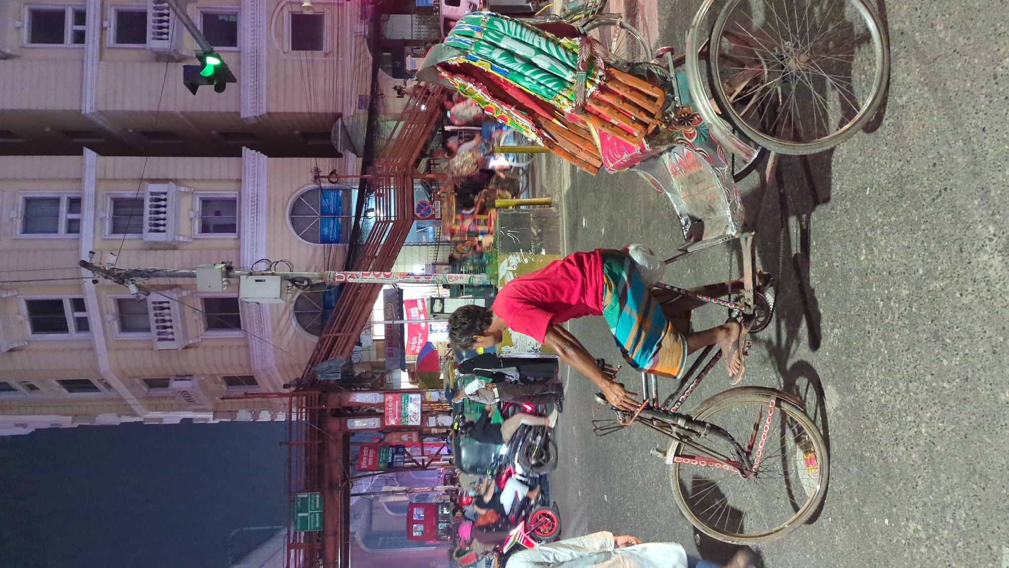 A man in a red shirt and striped shorts is operating a cycle rickshaw on a street in Dhaka, Bangladesh. The rickshaw's passenger seat is decorated with colorful cushions and fabric. Behind the rickshaw, a busy street with motorcycles, a red bus, and pedestrians is visible. Overhead, a dense network of electrical wires and signs crisscrosses the scene. A tall building with multiple windows and balconies stands to the left, with a green traffic light attached to it. The scene is illuminated by streetlights, and the pavement has a dark, textured surface.