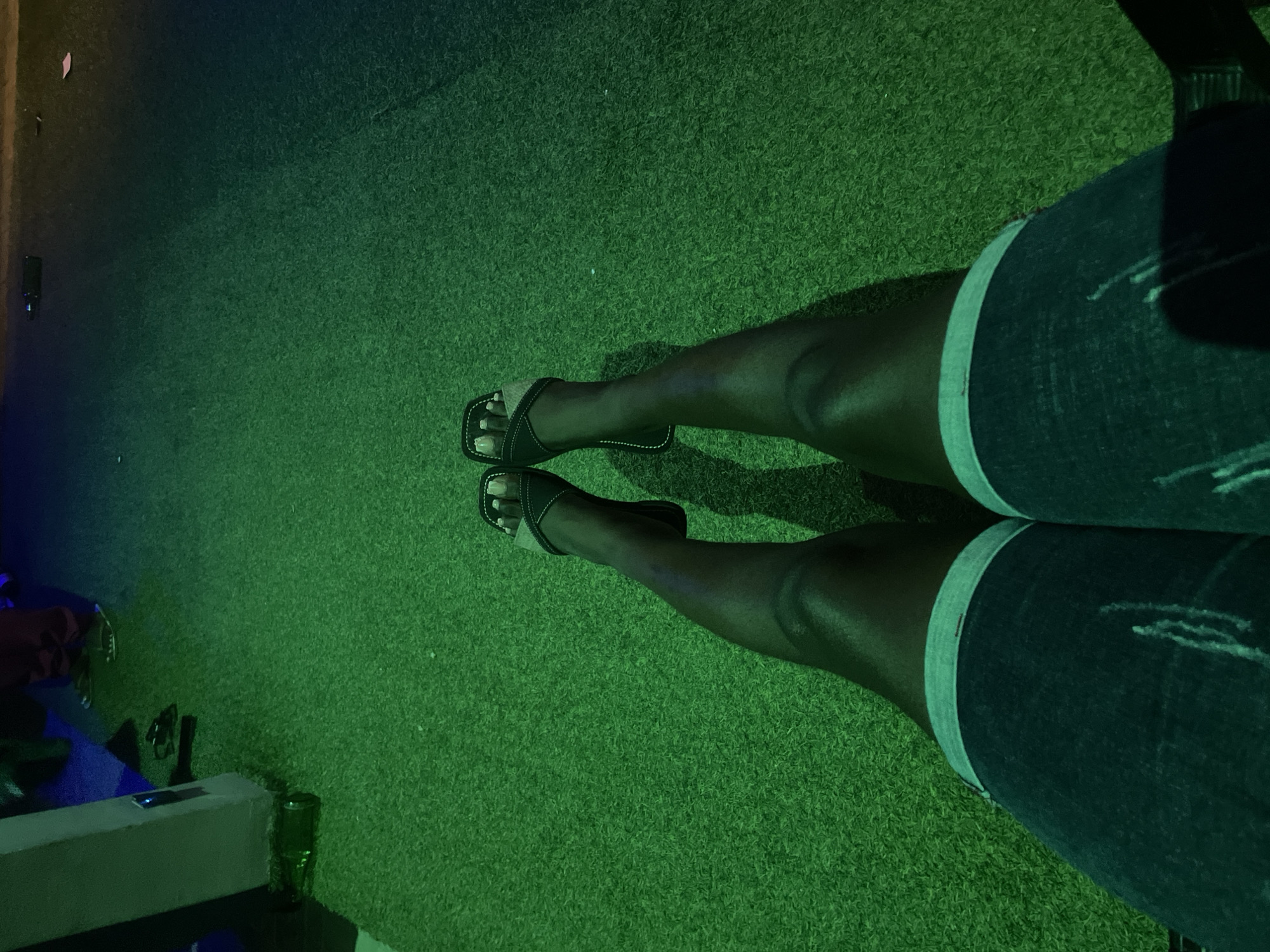 The image shows a person's legs and feet, wearing denim shorts and black sandals, resting on a green, artificial turf surface. The lighting is dim and cast in a greenish hue, with some areas transitioning to a darker, bluish-purple tone. In the background, a concrete pillar is visible on the left, with a glass bottle on the ground near its base. A person's legs and feet, wearing red pants and sandals, are also partially visible in the upper left background. The scene appears to be an outdoor setting, possibly a recreational area or an establishment with artificial turf. The location is identified as Kason, Nigeria.
