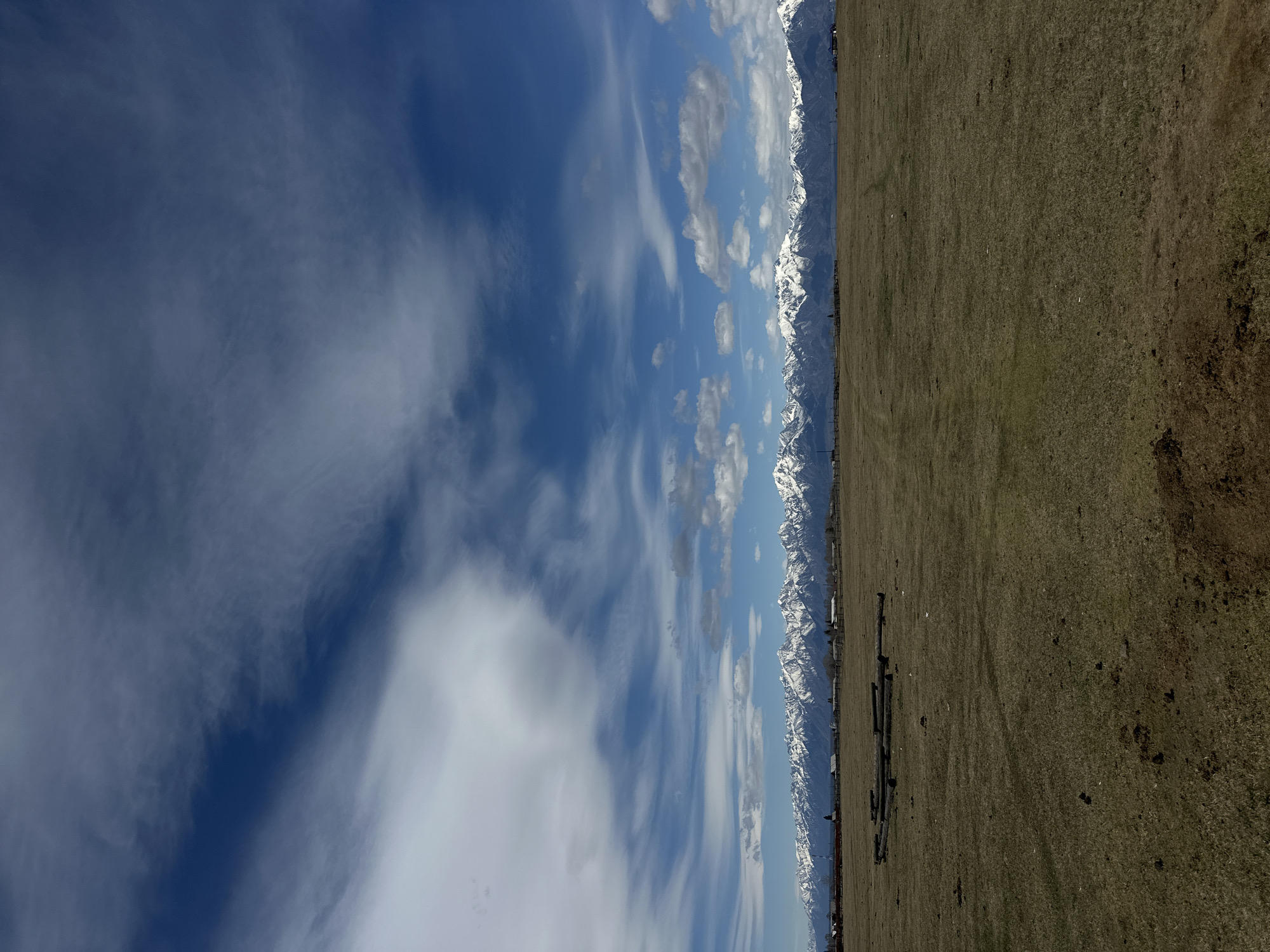 This image presents a broad horizontal landscape under a clear daytime sky, divided into three main sections: sky, distant mountains, and a foreground field.
The top portion features a vibrant blue sky dotted with scattered white clouds. These clouds range from wispy, translucent formations that streak across the expanse to more defined, cumulus-like patches.
Along the horizon, a prominent range of snow-capped mountains stretches across the middle ground. Their rugged peaks are covered in bright white snow, contrasting with the darker hues of the rock, indicating a high-altitude or cold climate region.
The lower half of the image is an expansive, dry field covered in brownish-green grass or dirt, suggesting a rural or undeveloped area. In the foreground, several dark, elongated objects, possibly pipes or logs, lie on the ground towards the left and center. Further left, near the base of the mountains, indistinct dark structures, possibly fencing or distant buildings, are visible.
The scene depicts fair weather during daylight hours, with no strong shadows to indicate early morning or late evening. No people or specific activities are present, highlighting the natural beauty and vastness of the environment. There is no visible text. This tranquil landscape is situated in Galbai, Russia.