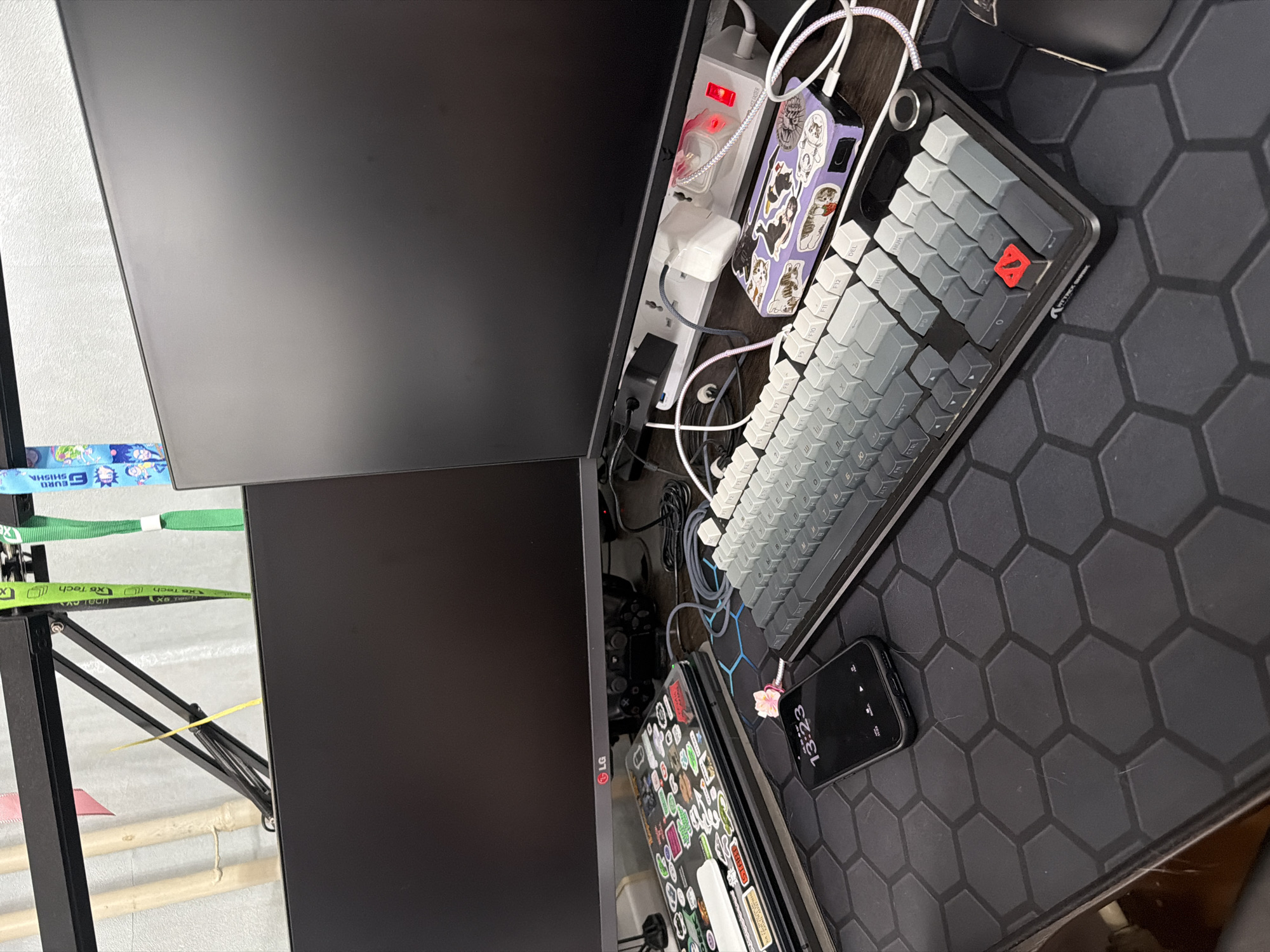 The image shows a desk setup with two computer monitors, a keyboard, a smartphone, and a power strip. The left monitor has an LG logo. A power strip is plugged into the wall and has several items plugged into it, including a white power adapter. A laptop is placed on a stand below the left monitor and is covered in stickers. A smartphone is on the desk displaying the time 13:23. A keyboard with grey and white keys is in front of the right monitor. There are several lanyards hanging from a shelf above the monitors. The environment appears to be indoors, likely an office or home setup. There are no people visible in the image. The image does not provide any information that would allow for identification of its location in the city of Chaikovsky, Russia.