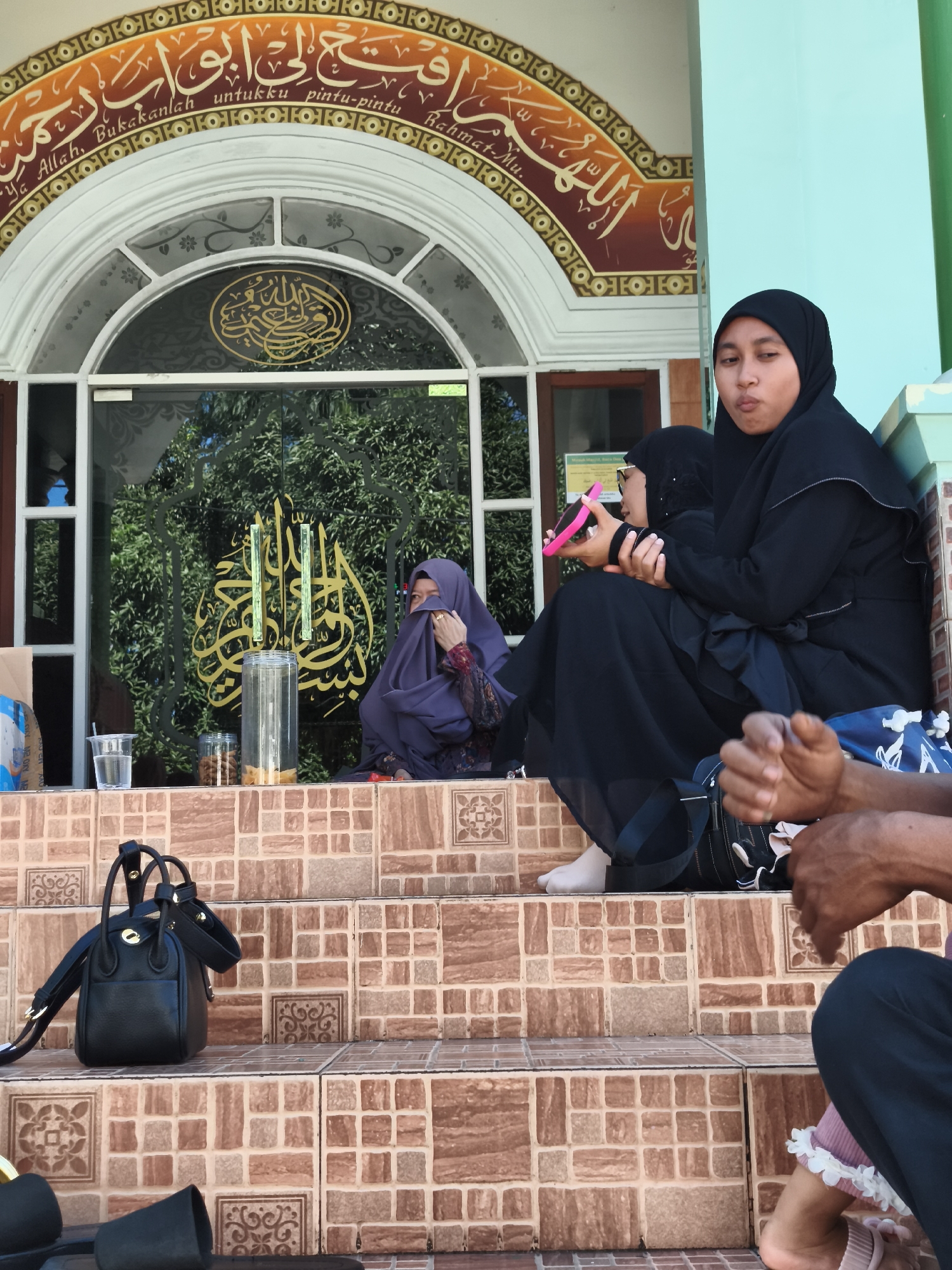 The image captures a scene outside a mosque in Tonasa, Indonesia. In the foreground, a woman wearing a black hijab and dress is seated on tiled steps, looking towards the camera with a surprised expression. Next to her, another person, partially visible, is holding a pink object. Further back, a person in a purple hijab is seated. The mosque entrance features glass doors with Arabic calligraphy in gold and decorative floral patterns. The steps are tiled with a mosaic of brown and beige squares, some with ornamental designs. A black handbag rests on the steps. In the background, lush green trees are visible through the glass doors. The overall atmosphere suggests a casual gathering or a moment of rest outside the place of worship.