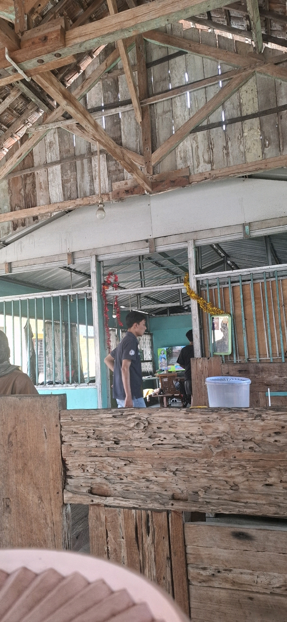 The image depicts an indoor scene, likely within a building featuring a rustic, open-air design. The prominent architectural elements include exposed wooden beams supporting a weathered roof structure made of corrugated metal and wooden planks, with visible gaps allowing sunlight to filter through. The walls are constructed from vertical wooden planks in various shades of gray and white, suggesting age and exposure to the elements.
In the foreground, a large, textured wooden beam occupies the bottom portion of the frame, its surface riddled with holes and imperfections. To the left, a person wearing a brown head covering and a brown garment is partially visible.
The mid-ground reveals a more defined area with teal-colored walls and a light blue ceiling. A metal grate structure with vertical bars runs along the wall, partially enclosing the space. A young man in a dark blue t-shirt and light blue jeans stands near the grate, facing away from the camera and looking towards the right. He has his left hand on his hip. Behind him, a television screen displays an image, and a person in a black shirt is visible on the far right. A yellow garland hangs from the grate on the right side, and a red garland is draped on the left side. A translucent blue plastic container is placed on a surface to the right.
In the background, another person is seen lying down on what appears to be a chair or a bench. The overall impression is a casual, possibly commercial setting, perhaps a cafe or a communal space. The image appears to be taken in Pabelan, Indonesia, given the context provided.