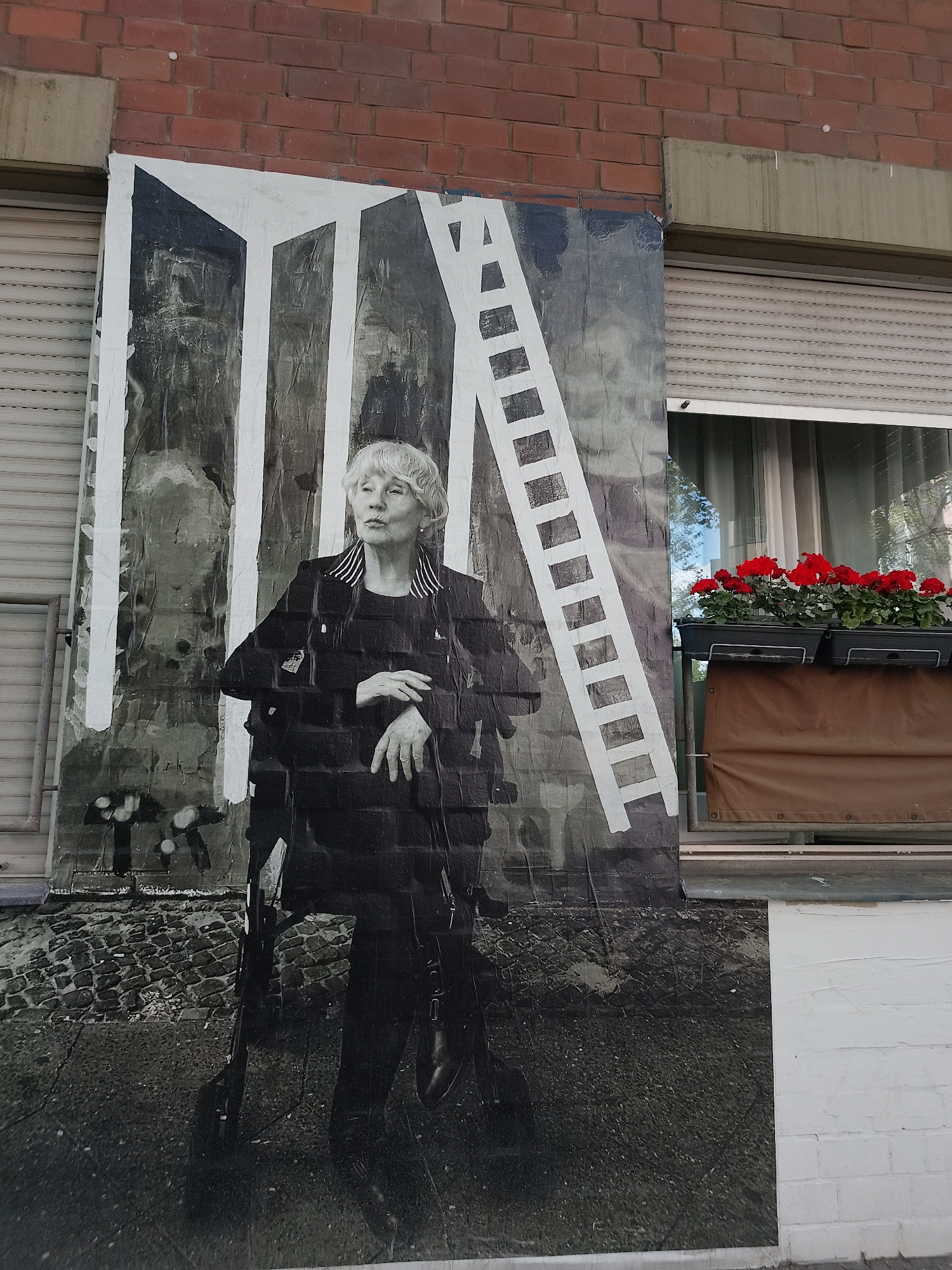 A large poster is affixed to the brick exterior of a building in an urban setting. The poster features a black and white image of an elderly woman with short white hair, wearing a dark outfit. She has her arms crossed and is looking off to the side. Behind her, a design with white stripes and a ladder-like structure is visible. The poster is partially peeling and has some marks. To the right of the poster, there's a window with closed blinds and a window box filled with red flowers. The bottom of the building is painted white. The ground in front of the poster is part of the printed image, depicting a street or pathway.