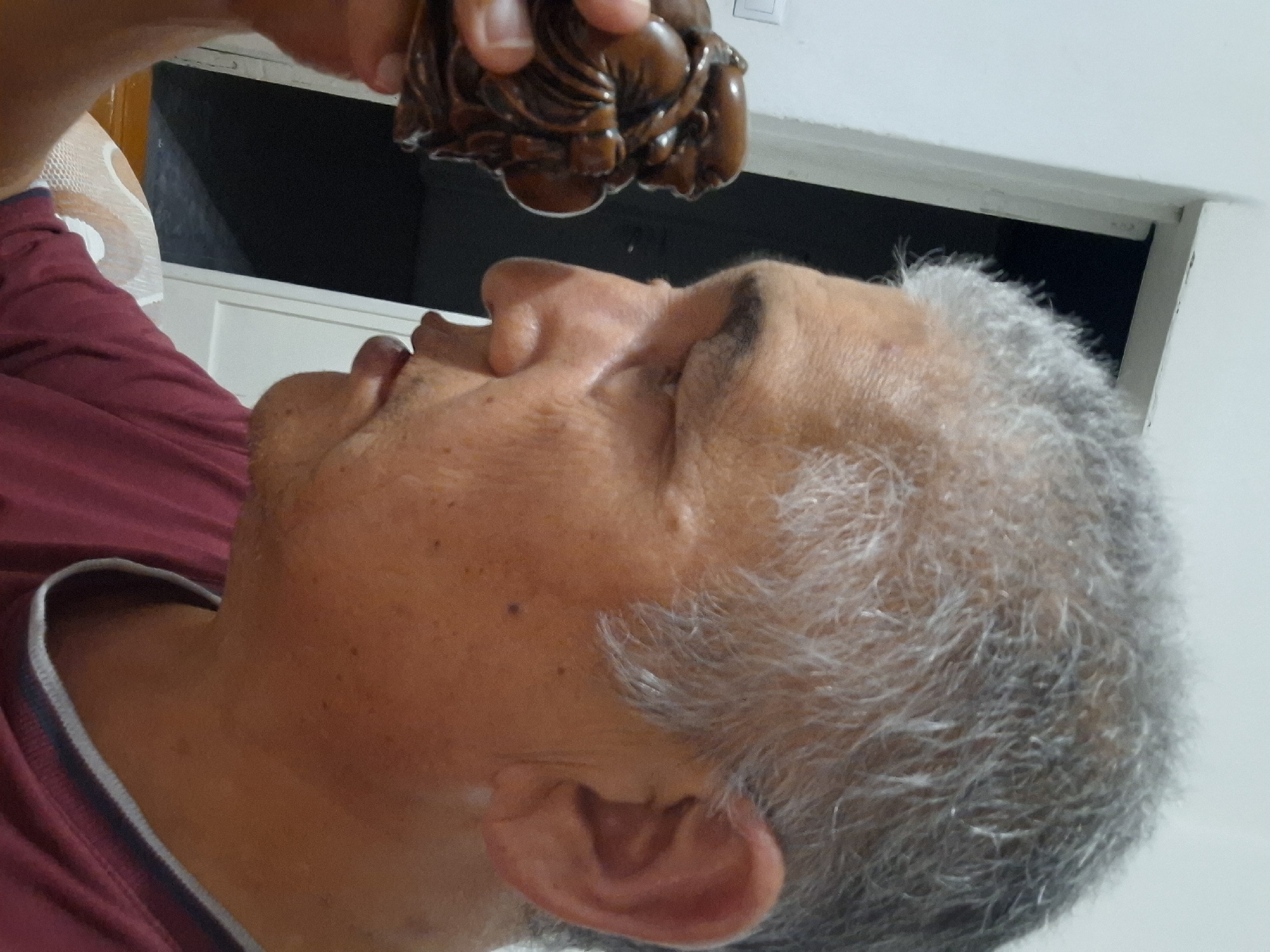 In a medium shot, an elderly man with graying hair is holding a small, dark brown, carved figurine of a plump, seated figure, possibly of a deity or a mythical character, near his face, looking at it closely. He is wearing a dark red shirt with a gray and dark blue trim around the neckline. The background shows a white wall with a doorway to a dark room and a light switch on the wall. The scene appears to be indoors, in a residential setting in Almaty, Kazakhstan, though no specific landmarks or street views are visible to confirm the exact location within the city.
