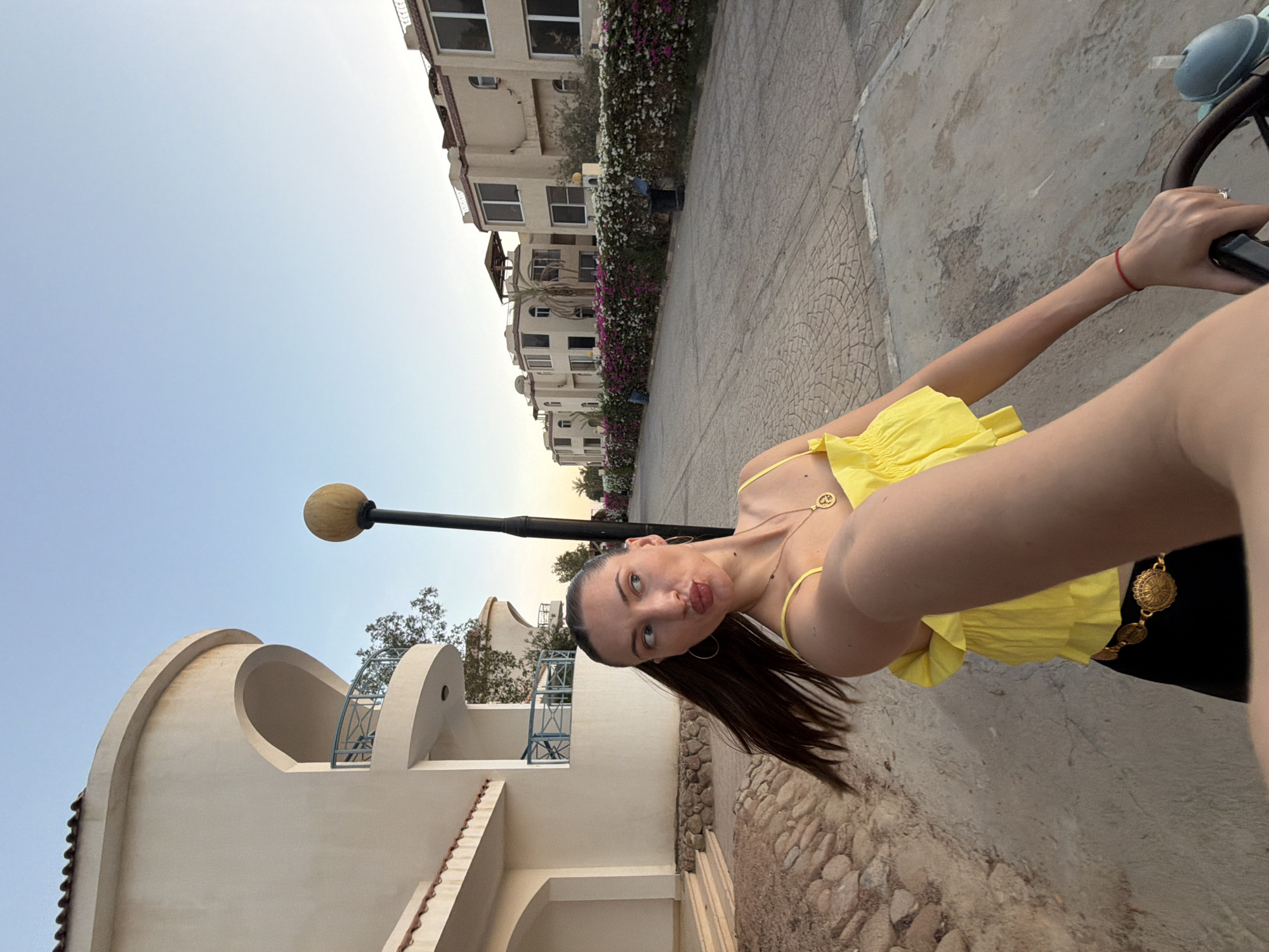 The image depicts a young woman in a yellow top and black skirt with a gold belt, posing for a selfie while sitting on a bicycle. She is wearing a necklace with a pendant and hoop earrings. Her hair is long and dark, tied in a ponytail. The background shows a street lined with buildings, a row of colorful flowers, and palm trees. The architecture suggests a resort area, possibly in Sharm El Sheikh, Egypt, with its characteristic pale-colored buildings and balconies. The street is paved with cobblestones. The lighting indicates it is late afternoon or early evening. The overall scene conveys a vacation or leisure setting.