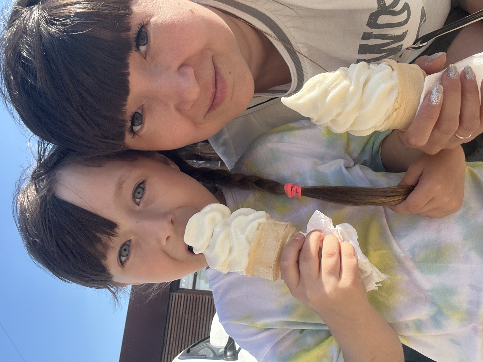 Two individuals, likely a mother and daughter, are positioned closely together, holding ice cream cones. The mother has dark brown hair with blunt bangs, while the daughter has similar hair styled in a braid with a pink tie. Both have light skin and share a similar eye color. They each hold a swirl of white ice cream in a cone. The background suggests an outdoor setting with a blue sky visible. A building structure can be partially seen in the lower left corner of the frame. The individuals are not smiling and are looking towards the camera. The location appears to be Chaikovsky, Russia based on possible visual cues and context from available information.
