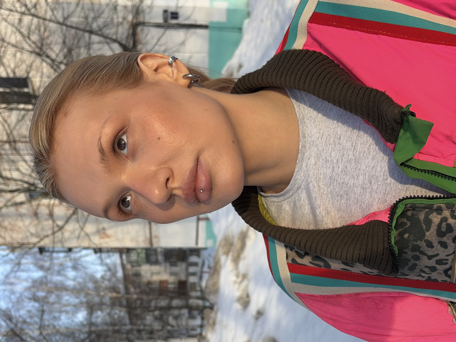 The image depicts a young adult female outdoors in Chaikovsky, Russia, during winter. She is shown from the chest up, positioned slightly to the right of the frame, gazing upwards and to her right. The female has blonde hair pulled back, a fair complexion, and light-colored eyes. Her lower lip is pierced with a silver labret stud. Her visible left ear features a silver hoop earring in the lobe and a silver ear cuff on the helix. She is wearing a grey crew-neck t-shirt beneath a vibrant, multi-colored jacket. The jacket is predominantly hot pink, featuring a dark brown ribbed collar. Vertical stripes in light blue, white, and red are visible on the jacket's shoulders. A leopard print pattern (brown and black) is apparent on an inner lining or trim, and green fabric elements are visible near the zipper. The background consists of a snowy ground, bare deciduous trees, and multi-story residential buildings with light-colored facades. One building further right has a light blue and white painted lower section. The scene is illuminated by natural sunlight, casting warm light on the subject's face.
