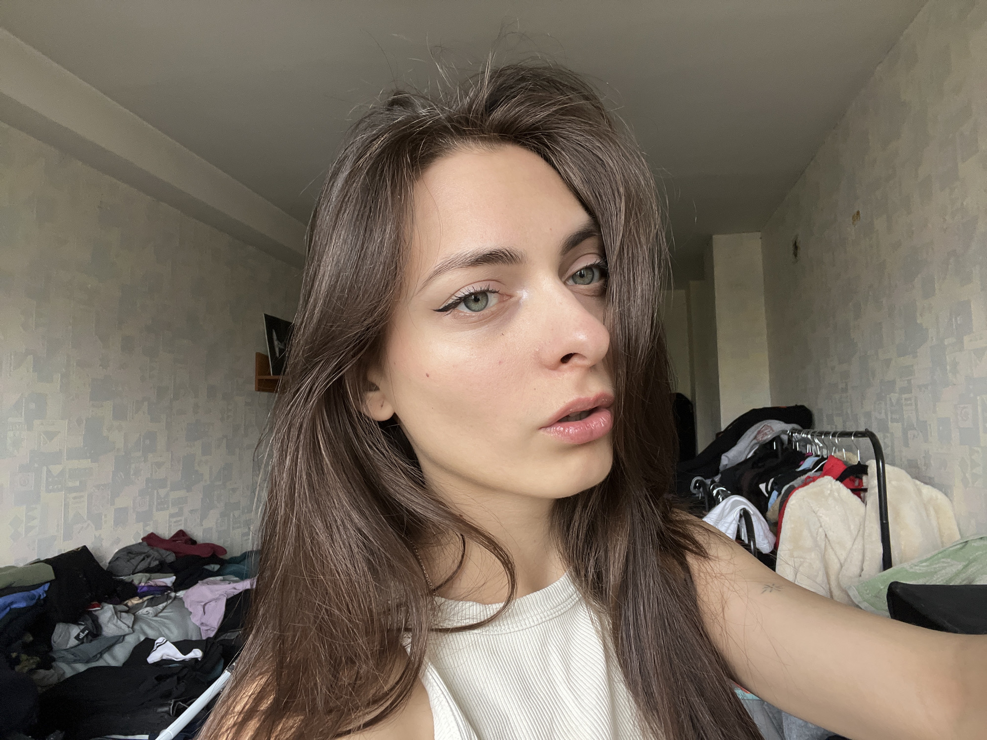 A young woman with long brown hair, fair skin, and green eyes is taking a selfie. She is wearing a white ribbed tank top and has eyeliner on. The background appears to be a bedroom with a bed covered in clothes on the left and a clothing rack with assorted items on the right. The wallpaper is patterned in a light grey and white design. Given the details in the image, no information is present indicating a specific location in Krasnoyarsk, Russia.