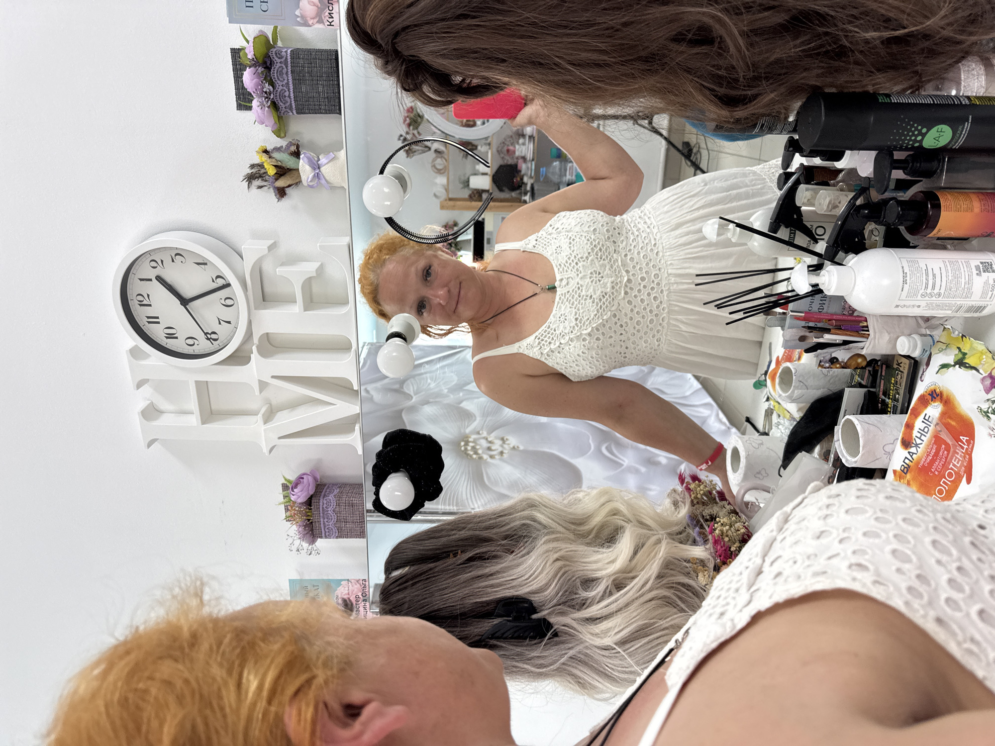 The image depicts a scene inside a beauty salon, possibly a wig shop, in Chaikovsky, Russia. The primary focus is on a woman with red hair standing in front of a mirror, wearing a white lace dress and a necklace. She is holding a pink object, possibly a phone or a small tool, in her right hand and looking towards the viewer. In the mirror's reflection, another woman with blonde hair, seen from the back and side, is also present. The salon environment is indicated by various objects on shelves and a counter, including decorative items, bottles of hair products, brushes, and a clock on the wall that reads approximately 11:55. The overall atmosphere suggests a professional or personal grooming setting.