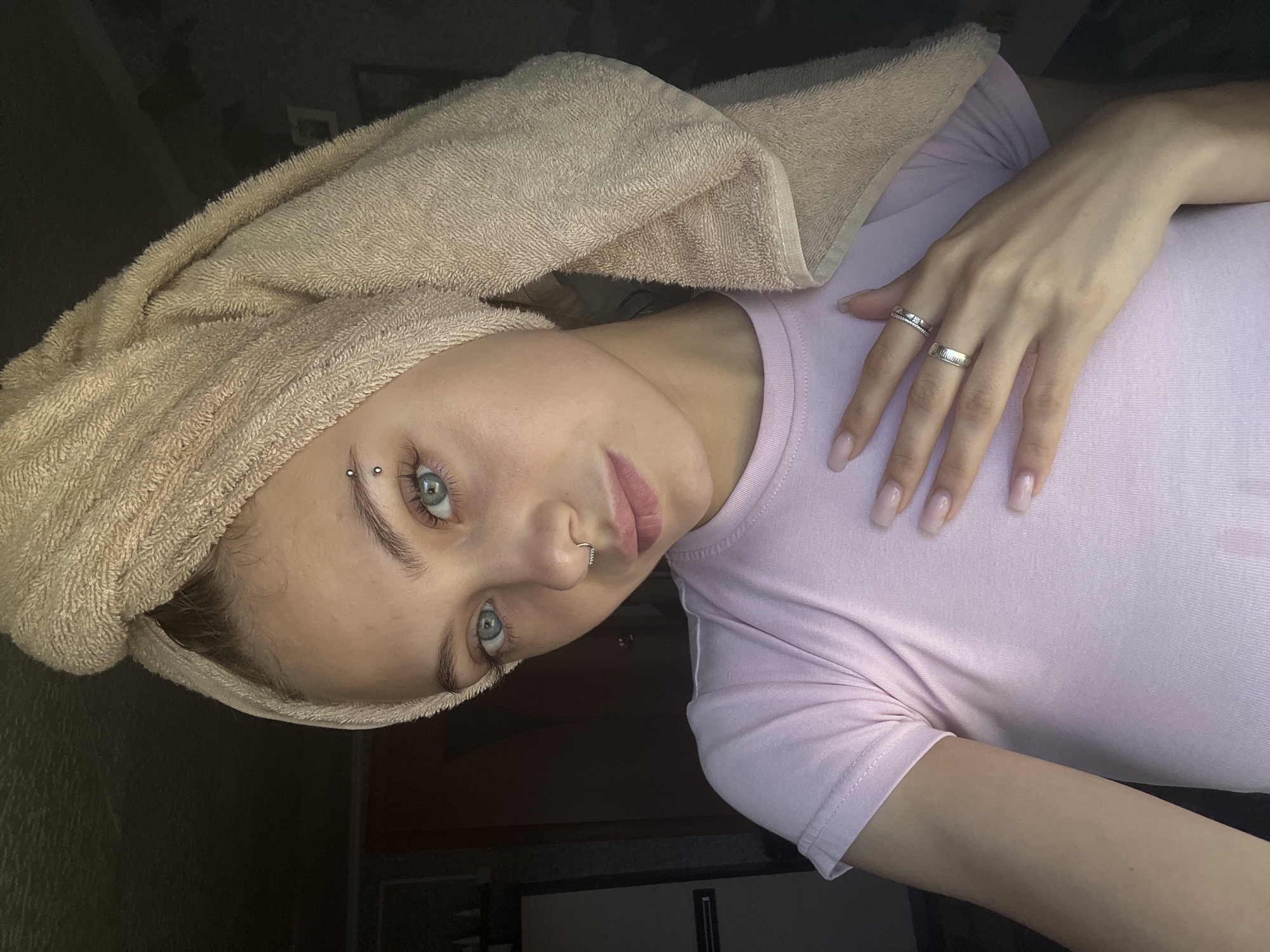 The image shows a young woman with blue eyes and a light brown towel wrapped around her head. She is wearing a pink t-shirt and has a nose ring and an eyebrow piercing. Her left hand is resting on her chest, displaying two rings on her fingers. The background is dimly lit with some furniture visible, suggesting an indoor setting, possibly a bedroom or bathroom. The image does not provide any specific information or visual cues that would allow for the determination of its location within the city of Fokino, Russia.