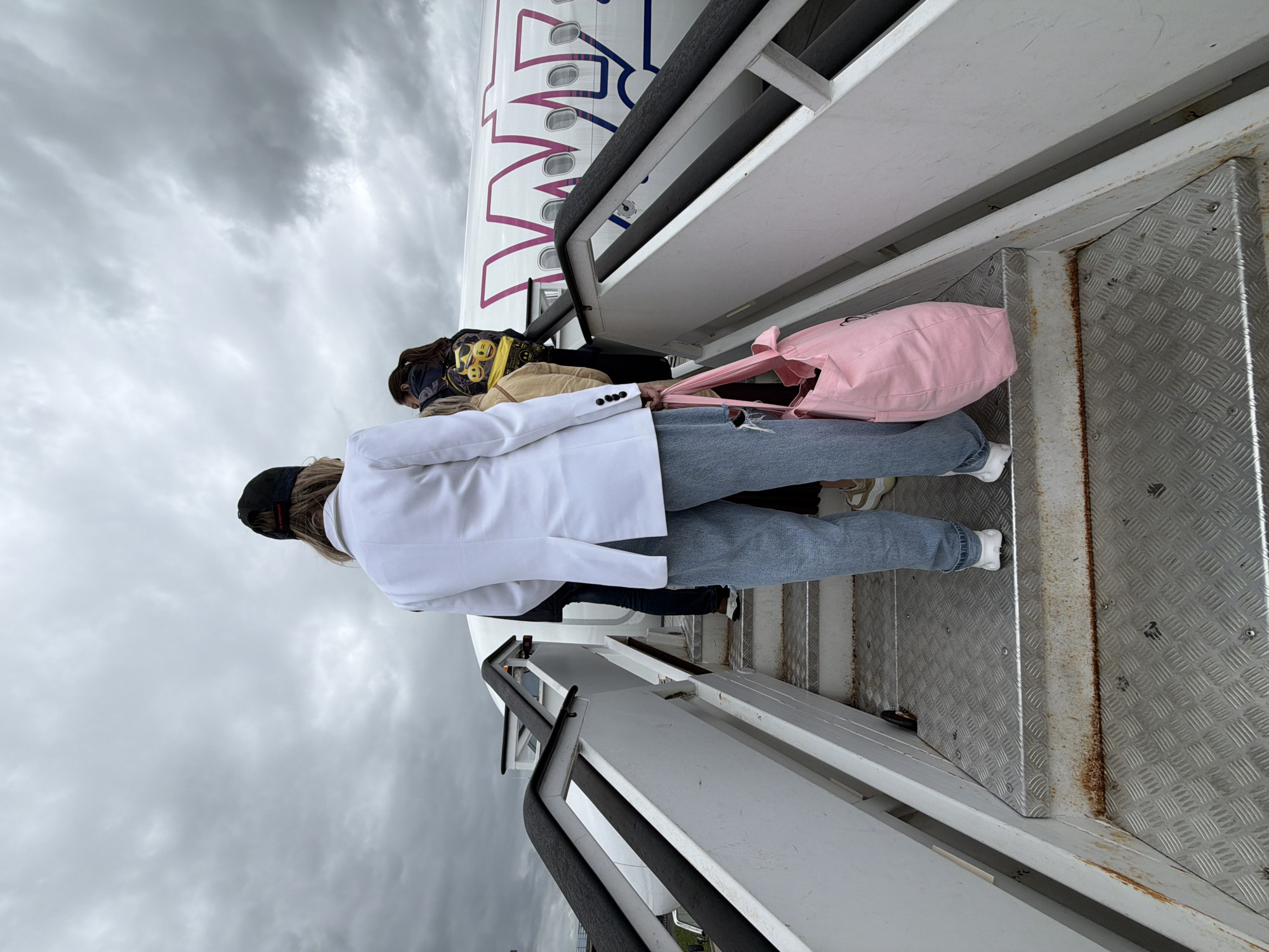 The image depicts a scene of passengers boarding a commercial airplane via a mobile staircase under an overcast sky. The setting is likely an airport, specifically Tillé, France (Beauvais–Tillé Airport), given the context.
The perspective is from behind a person ascending the metal airstairs. This individual is wearing a white long-sleeved top or blazer, light blue jeans, white sneakers, and a black baseball cap worn backwards. They are holding a light pink tote bag in their right hand. Behind them, another person is partially visible, wearing a tan or beige top and a dark jacket with yellow patterns. Further up the stairs, another passenger can be faintly seen.
The metal staircase features diamond-plate steps, some showing signs of rust and wear, and white side railings with black handrails. The airplane's fuselage is white with a distinctive purple/pink logo and lettering. Parts of the airline's name, resembling "WIZZ", are visible, strongly suggesting the aircraft belongs to Wizz Air. Several airplane windows are discernible along the side of the fuselage. The sky is completely covered with thick, grey clouds, indicating a cloudy daytime. The overall activity is people boarding an aircraft.