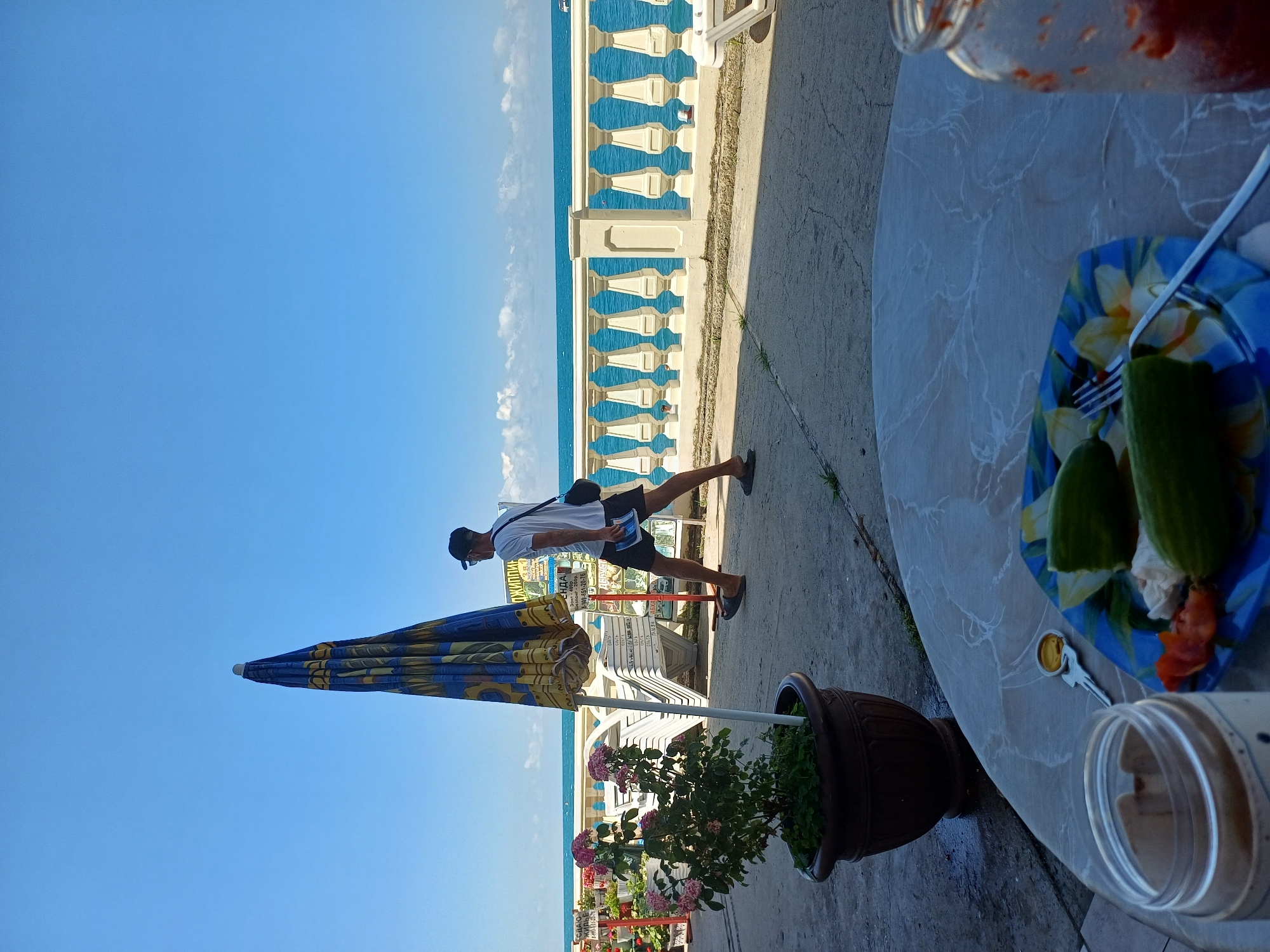 A man wearing a white t-shirt, black shorts, a black baseball cap, and a black crossbody bag is walking along a concrete path beside a white and blue balustrade overlooking the turquoise sea. The sky is a clear, bright blue, suggesting a sunny day. In the foreground, a colorful beach umbrella with a blue and yellow pattern is partially closed. To the right of the umbrella, a potted plant with pink flowers adds a touch of color to the scene. Further to the right, a round table is set with a blue plate holding what appears to be food, a fork, and a jar with a red substance. A second potted plant, this one filled with green foliage, sits near the table. The scene appears to be a casual outdoor setting, likely a promenade or a cafe by the sea, possibly in Sochi, Russia, given the context. The overall mood is relaxed and summery. There is some Cyrillic text visible on a sign behind the man, but it is not clearly legible.