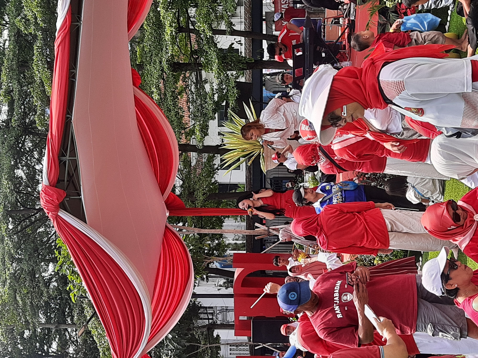 In Bandung City, Indonesia, a crowd gathers under a red and white draped canopy, likely for a festive event. Two female singers perform on a stage, one in a red dress, the other in a white jumpsuit, both holding microphones. A keyboardist plays an instrument to their right. Among the attendees, many are wearing red, and some are adorned with Indonesian flags or colors, suggesting a national celebration. A man in a red shirt with "RANTJABADAK" printed on it is visible in the foreground, holding a phone. The scene is set outdoors with lush green trees in the background.
