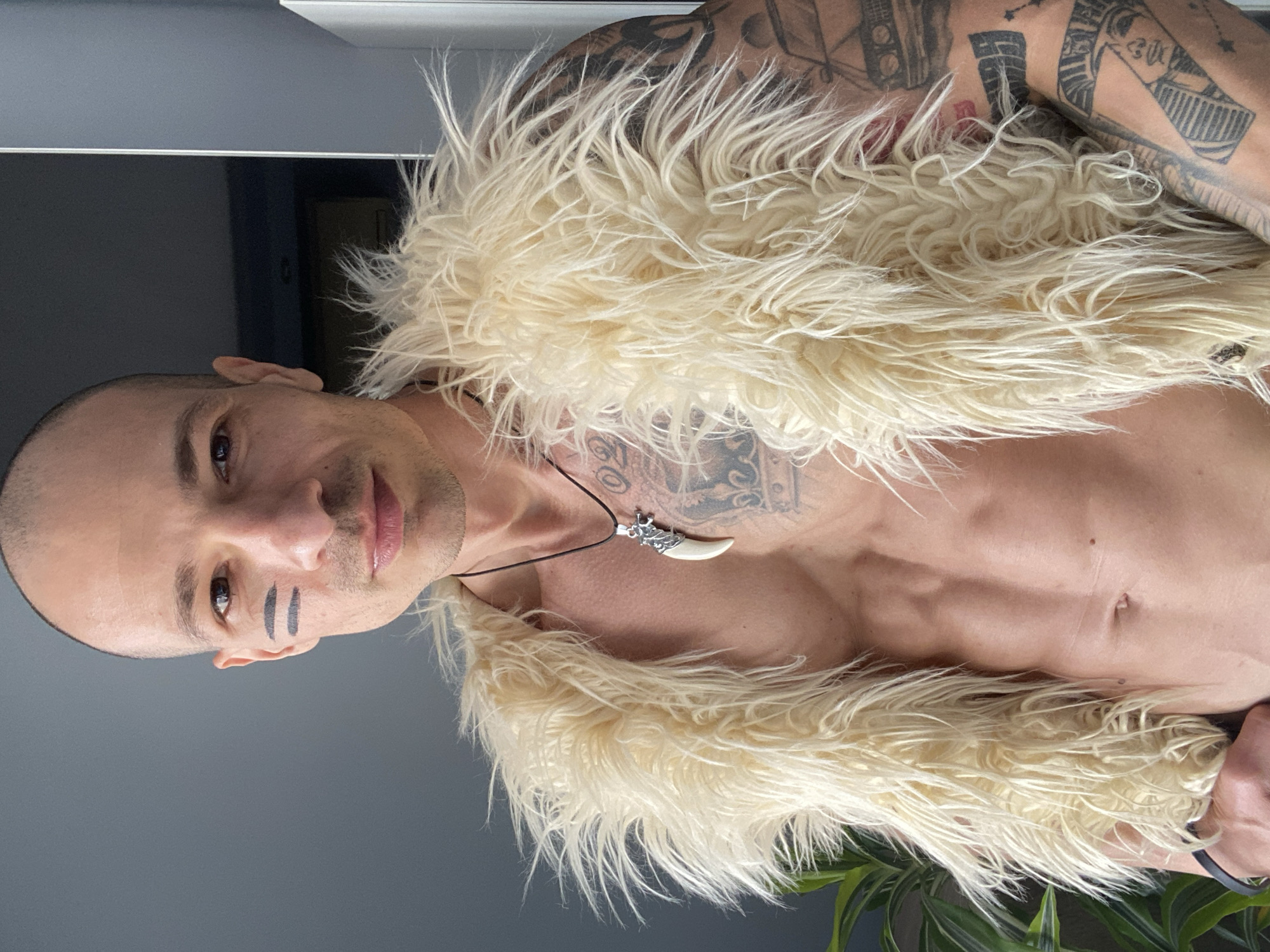 A man poses indoors. He has a shaved head and fair skin with tattoos on his upper chest and arms. Two black stripes are drawn under one eye. He is wearing a beige, shaggy fur vest and a necklace with a tooth-shaped pendant. The background is a gray wall with a hint of a door frame. A green potted plant is visible at the bottom of the frame. There is no information in the image that directly indicates that the location is Pleven, Bulgaria.
