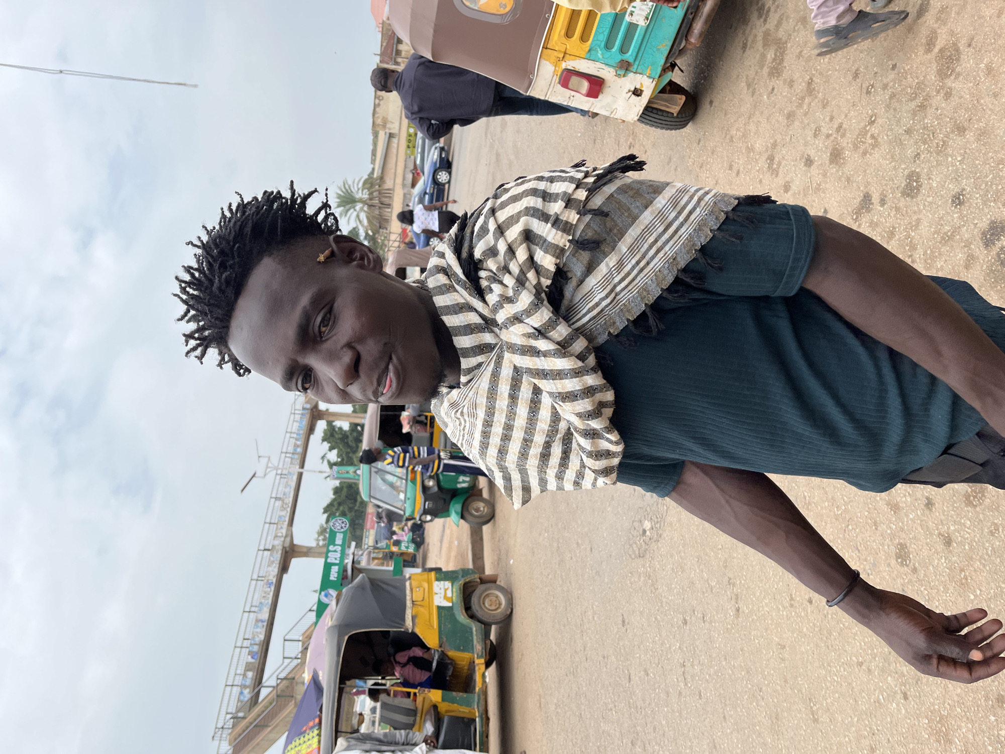 The photograph captures a medium shot of a young Black man with dreadlocks, wearing a dark green ribbed t-shirt and a horizontally striped scarf draped around his shoulders. He is standing outdoors on a paved surface, possibly a street or market area. In the background, several auto-rickshaws, commonly known as "keke napep," are visible, some appearing to be in motion or parked. One auto-rickshaw in the foreground has a predominantly yellow and green color scheme. Further in the background, a pedestrian overpass with stairs is present, and beyond that, buildings and trees are discernible. The sky is overcast with clouds. The overall scene suggests an urban setting in Nigeria.
